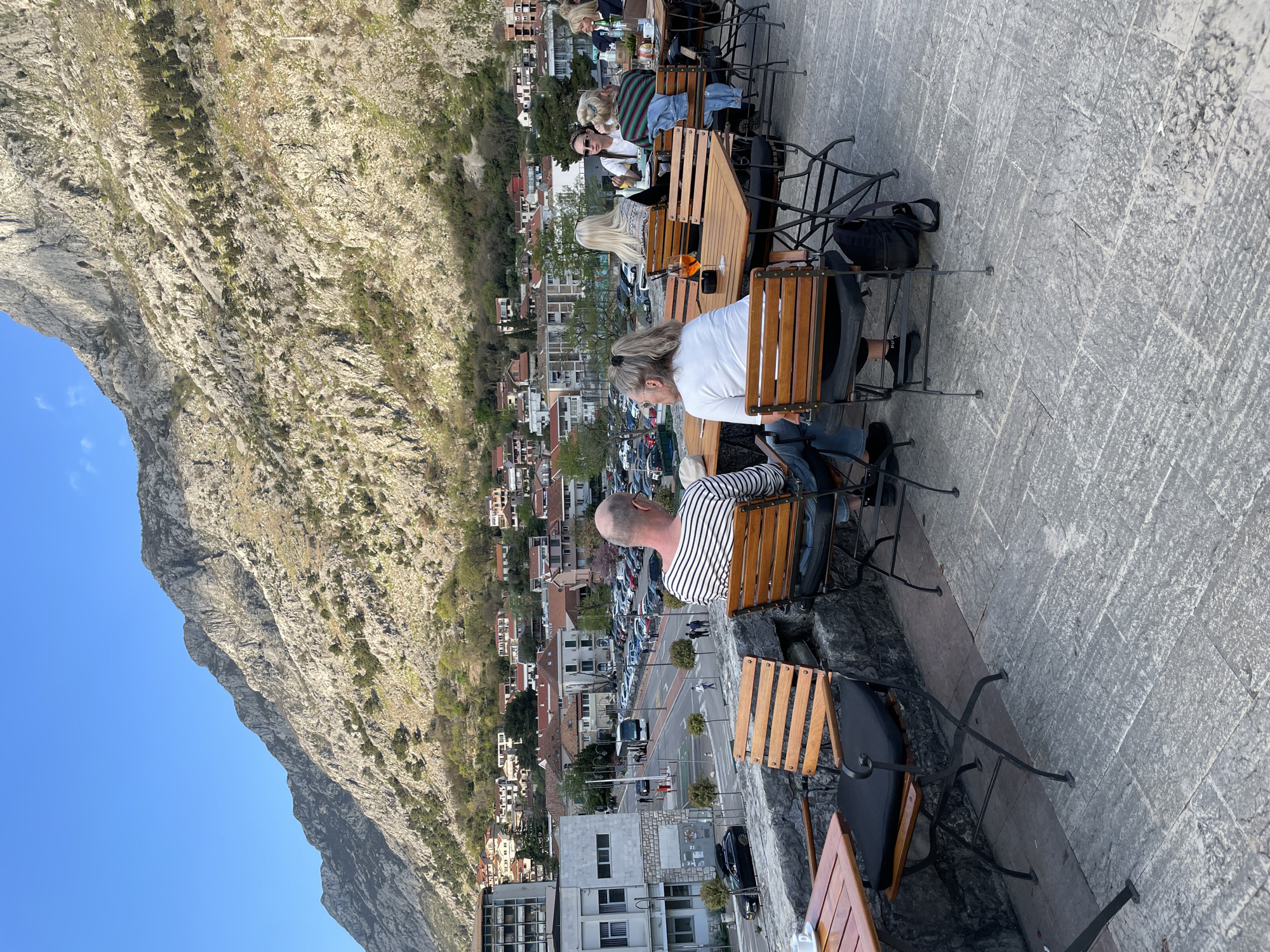 The image shows a restaurant scene in Kotor, Montenegro, with patrons seated at wooden tables and chairs on a stone terrace overlooking the city and a large mountain. Several people are visible, including a man in a striped shirt and a woman in a white shirt, sitting at one of the tables. Other patrons are seated at tables nearby. In the background, the city of Kotor is nestled among buildings and trees, with cars parked along the streets. A large, rocky mountain looms over the city, filling a significant portion of the frame. The sky is clear and blue.