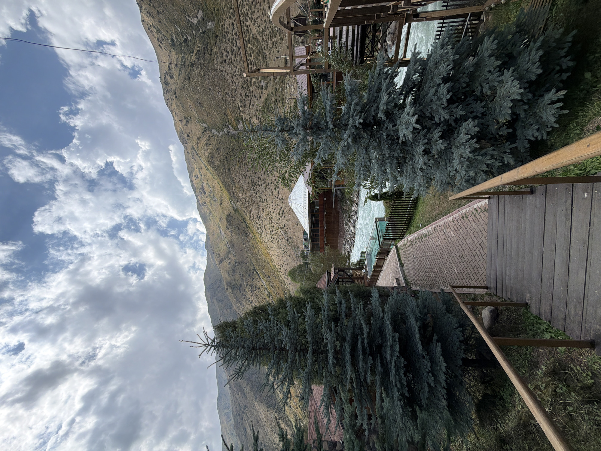 A wide shot captures a scenic view of a mountain resort along a rushing river under a cloudy sky. In the foreground, a wooden staircase with a handrail descends from the viewer's perspective. To the left and right of the stairs are large evergreen trees. A brick pathway leads from the bottom of the stairs to the left, curving around a small, fenced-in pool and continuing towards a building with a conical roof.
The river flows rapidly from left to right across the mid-ground, bordered by a wooden fence and more evergreen trees. Beyond the river, wooden structures with visible railings and roofs are situated. In the background, a steep, barren mountain slopes upwards, its surface textured with dry vegetation and scattered rocks. The sky is filled with dramatic, textured clouds against a pale blue backdrop. The scene is serene and natural, suggesting a location for outdoor recreation or relaxation.