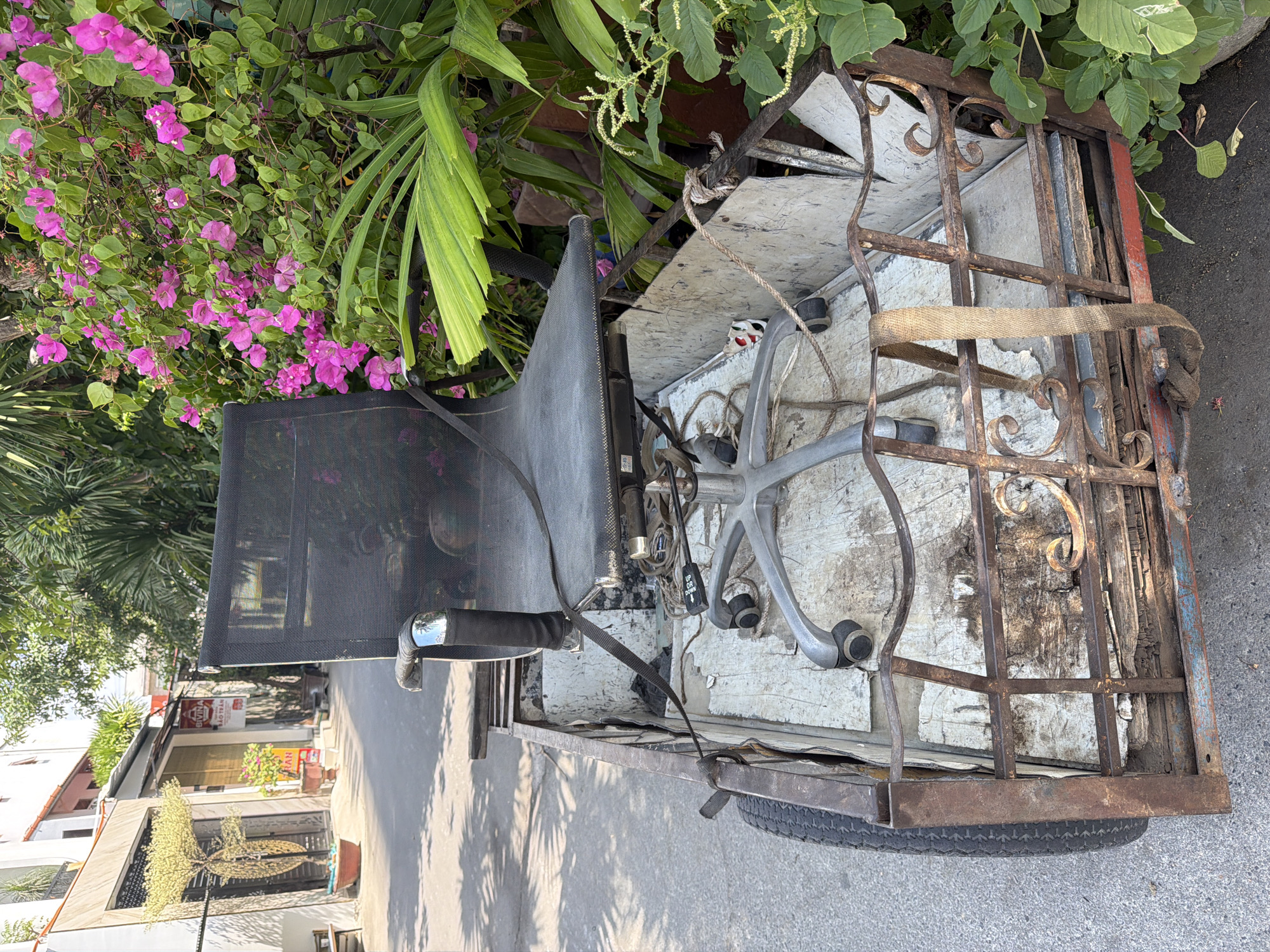 A black office chair with a metal base and wheels is secured with a tan strap inside a rusty metal cart with a single wheel. The cart is positioned on a paved surface, likely a street or alleyway, in Nha Trang, Vietnam. The background shows a building with a closed doorway and a small, light-colored tree in a pot. To the right of the cart is a dense green bush with vibrant pink flowers. The lighting suggests it is daytime, with shadows cast on the ground.