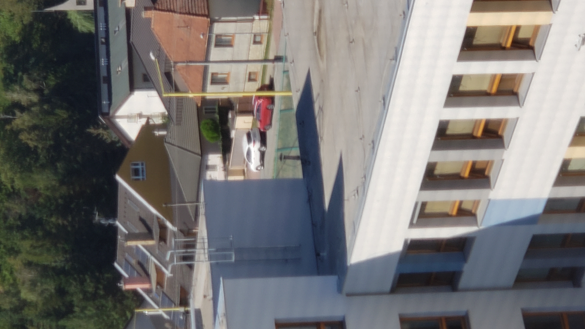 The image depicts a view of a residential area in Prešov, Slovakia, likely from a higher vantage point, looking down onto rooftops and the side of a building. In the foreground, the white facade of a modern building with numerous rectangular windows is visible. The windows have a light brown frame and appear to be tinted. The roof of this building is flat and gray, with visible mounting points for what could be solar panels or other rooftop equipment.
Further in the background, several houses with pitched roofs are visible, covered in grey and brown shingles. One house has a yellow facade and a prominent chimney. Several cars are parked on a street or driveway between the houses. A white SUV and a red hatchback are clearly identifiable. A tall yellow pole, possibly a lamppost, stands near the red car. Beyond the houses, a dense line of trees forms a dark green backdrop against a clear sky. The overall impression is of a sunny day in a suburban or semi-urban setting. The specific location within Prešov cannot be definitively determined from the visual information provided.