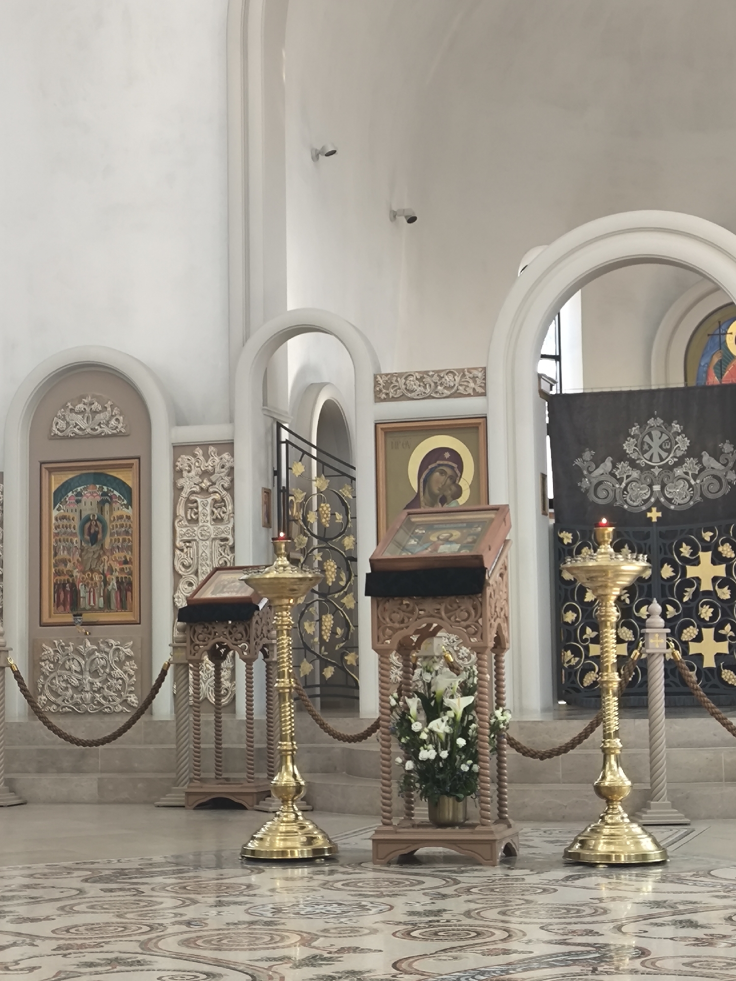 The image depicts the interior of a church, likely located in Strasbourg, France, given the architectural style. The scene features three arched entrances, each containing religious artwork and decorative elements. The arches are situated in a staggered arrangement, creating depth.
In the foreground, there are two ornate golden candelabras, each with a lit candle. They are positioned on either side of a wooden lectern, which holds a book. The lectern is adorned with a floral arrangement of white lilies. A rope barrier is strung between the candelabras and lecterns, delineating the space.
The background reveals more religious artwork, including icons framed in gold. The floor is covered with a patterned mosaic. The overall atmosphere suggests a place of worship with a focus on religious iconography and decoration. Two security cameras are on the wall near the ceiling.