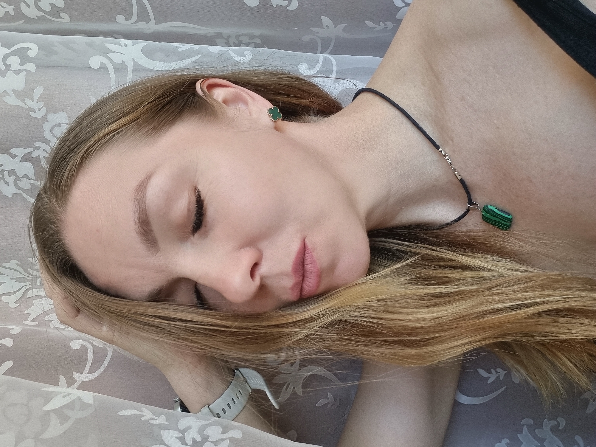 A blonde woman is lying down, eyes closed, on a surface with a white patterned fabric. She is wearing green four-leaf clover earrings and a black cord necklace with a green malachite pendant. A white watch with a perforated band is visible on her wrist. The location is Chaikovsky, Russia.