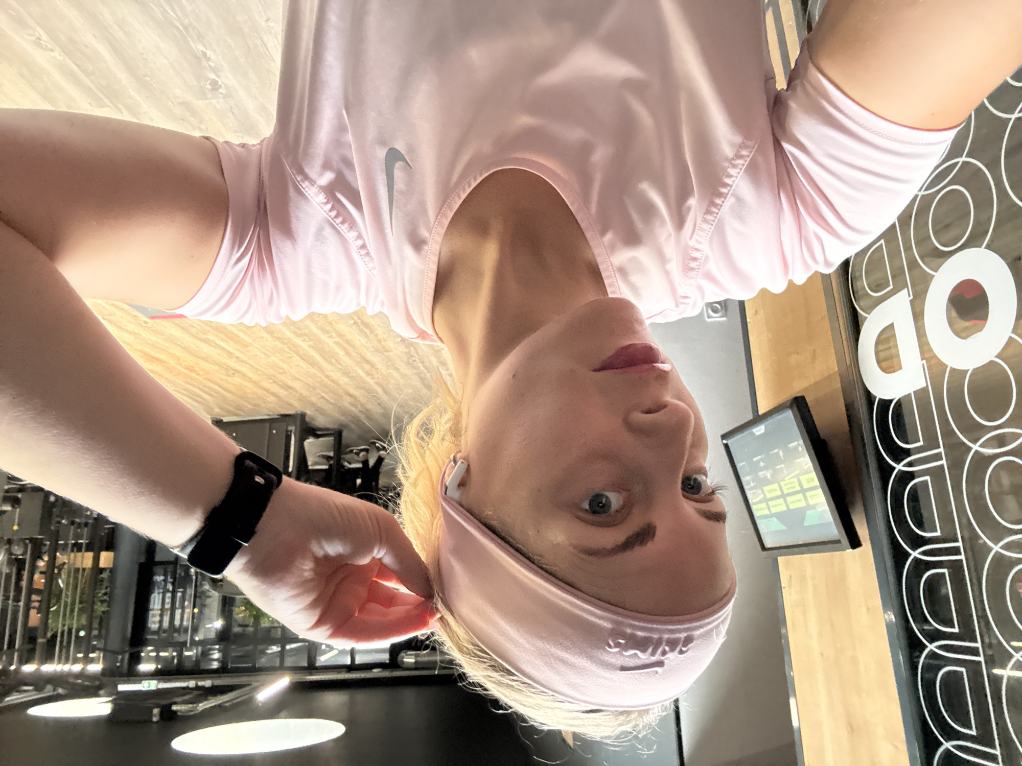 The image shows a selfie of a fair-skinned individual with blond hair, wearing a pink headband and pink athletic shirt. The person has a black smartwatch on their left wrist. The background appears to be a gym or fitness center with equipment and design elements. A digital display screen and a decorative wall panel with circular cutouts are visible. The gym appears to be located in Paris, France based on a review of available visual cues.