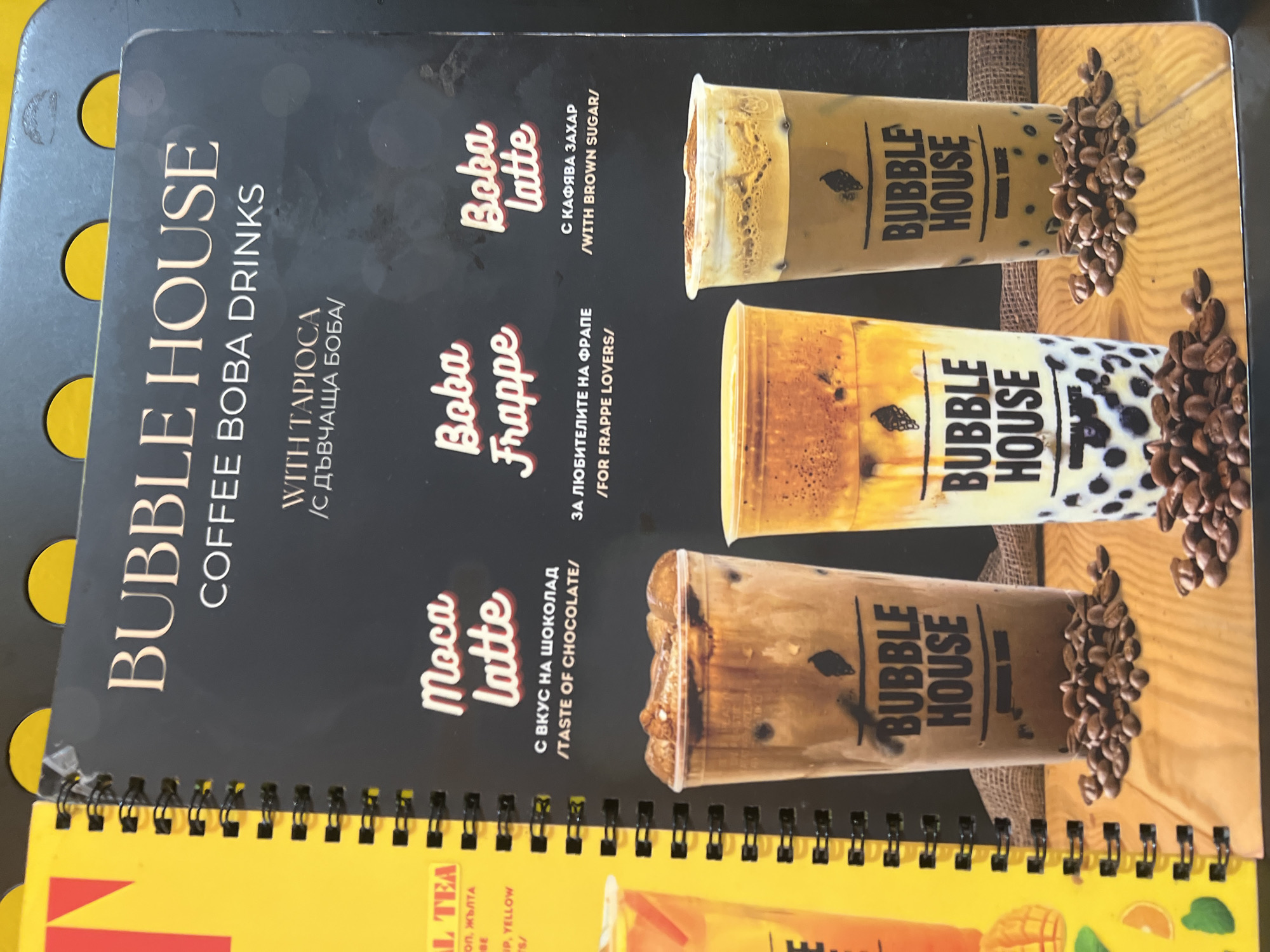This is a close-up shot of a menu page from "Bubble House Coffee Boba Drinks," likely from Plovdiv, Bulgaria, given the bilingual text. The dark gray menu displays three different boba beverages in clear plastic cups, arranged vertically on the right side against a light brown, textured background with scattered coffee beans. Each cup features the "BUBBLE HOUSE" logo.
The drinks listed are:
1. **Moca Latte**: Described in both Bulgarian ("С ВКУС НА ШОКОЛАД") and English ("TASTE OF CHOCOLATE").
2. **Boba Frappe**: Indicated for "FOR FRAPPE LOVERS" ("ЗА ЛЮБИТЕЛИТЕ НА ФРАПЕ").
3. **Boba latte**: Highlighted with "WITH BROWN SUGAR" ("С КАФЯВА ЗАХАР").
The menu prominently states that the drinks are "WITH TAPIOCA" ("С ДЪВЧАЩА БОБА"). The image is slightly rotated, with the menu's content oriented for landscape viewing. The top menu page is spiral-bound to a yellow page below it, which has partially visible text including "L TEA" and "YELLOW." The lighting is bright, clearly showcasing the layers and textures of the boba drinks and the surrounding coffee beans.