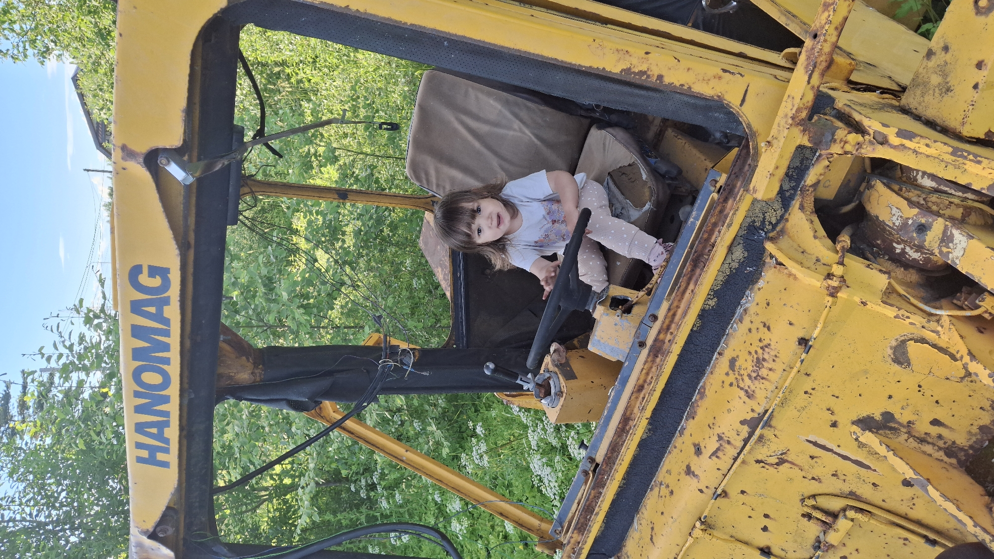 A young child, likely a toddler, is seated in the driver's seat of a weathered yellow construction vehicle. The child is wearing a white t-shirt with a floral design and light-colored pants with small polka dots. They have dark hair and bangs and are looking towards the camera with a slight smile. The child's hands are on the steering wheel, giving the impression they are operating the machine.
The construction vehicle appears old and rusty, with peeling yellow paint and visible metal wear. The interior of the cabin is visible, including a worn brown seat and various mechanical components, such as levers and the steering column. The vehicle is positioned outdoors, surrounded by lush green foliage, suggesting a rural or overgrown setting. The bright sunlight indicates it is daytime.
Visible text on the side of the vehicle reads "HANOMAG" in large blue letters. The overall scene evokes a sense of childhood exploration and imagination, with the child taking on the role of a builder or operator. The location context of Innolovo, Russia, places this scene within a specific geographical area, though the image itself does not contain explicit visual cues to pinpoint the exact location beyond general rural characteristics.