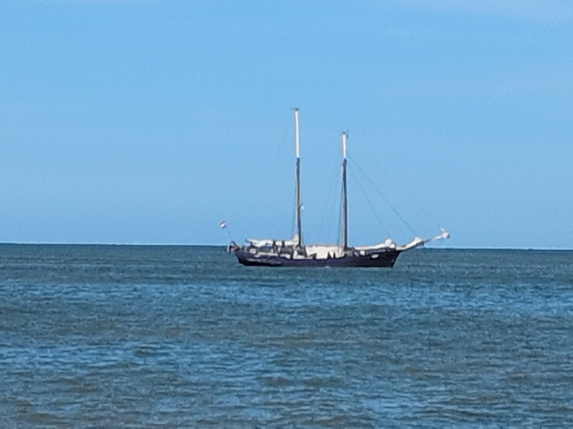 A dark-hulled sailing vessel with two masts is anchored on a calm sea under a clear blue sky. The ship has white superstructure and rigging. A small flag, possibly a national ensign, is visible on the bow. The water is a deep blue-green, with subtle ripples indicating a gentle breeze. The image does not provide sufficient detail to identify the specific location within Kühlungsborn, Germany, beyond it being a maritime scene. No people or specific actions are discernible.