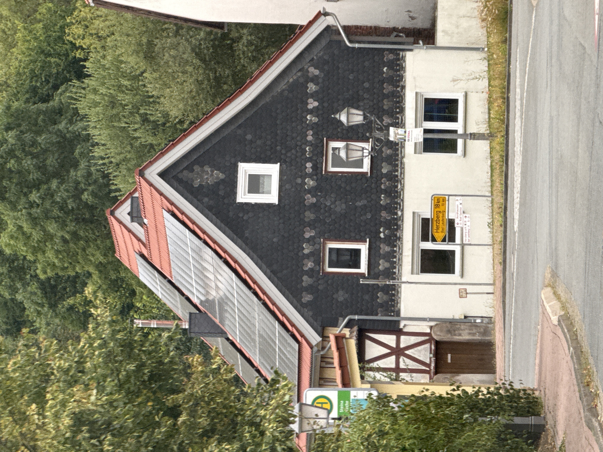 A residential house with a dark, patterned facade and a red-tiled roof featuring solar panels is shown in Bad Sachsa, Germany. The house has multiple windows, some of which are white-framed. A wooden door is visible on the side of the house. A street lamp with two light fixtures stands in front of the house, with a yellow directional signpost nearby indicating "Herzberg 18 km" and "Bad Lauterberg 10 km". A green and yellow street sign with a yellow circle and the letter "H" is mounted on a pole. Trees and foliage surround the property, and a road with a sidewalk is in the foreground. The sky is overcast, suggesting a cloudy day. There are no people visible in the image.