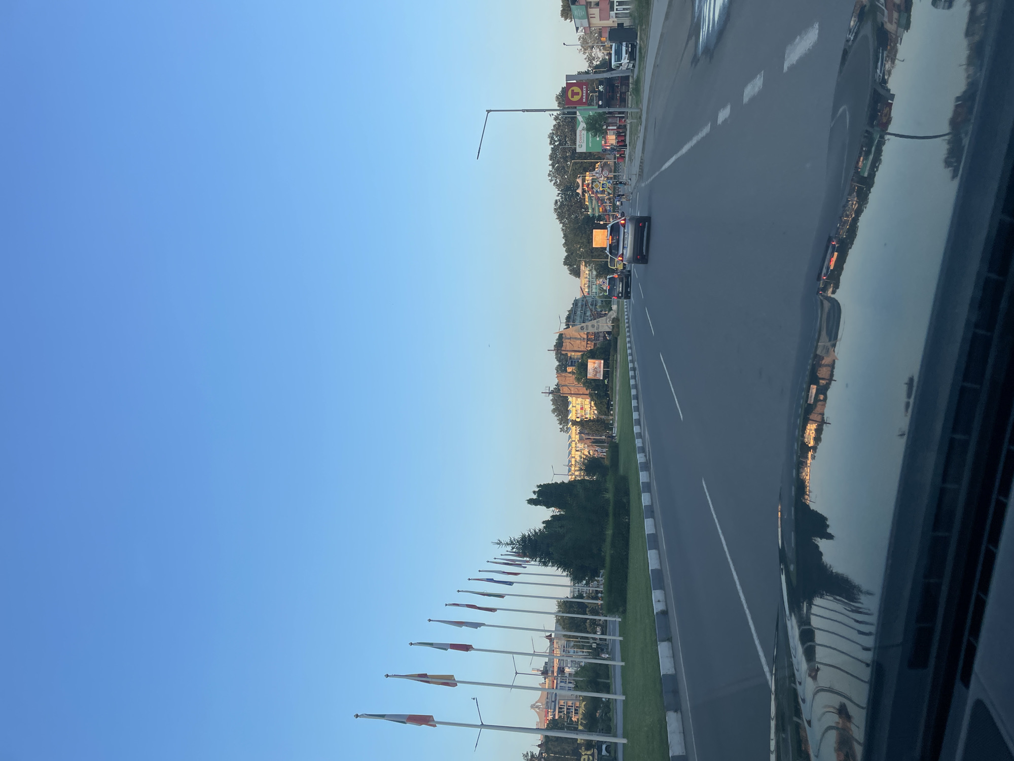 A wide, multi-lane asphalt road runs through a city on a clear, blue-sky day. The perspective is from a vehicle, looking forward. On the left side of the road, a grassy median separates the lanes and is lined with a curb and white and gray concrete barriers. Several tall flagpoles stand in the median, each flying a flag, many of which are colorful. Beyond the median are buildings and trees. On the right side of the road, there are more buildings, including what appears to be a gas station with a red and yellow sign and a "T MARKET" sign. Several cars are driving on the road in the same direction as the viewer. The hood of the vehicle the photo is taken from is visible in the foreground, reflecting the sky and buildings. The scene suggests an urban environment in Nesebar, Bulgaria.