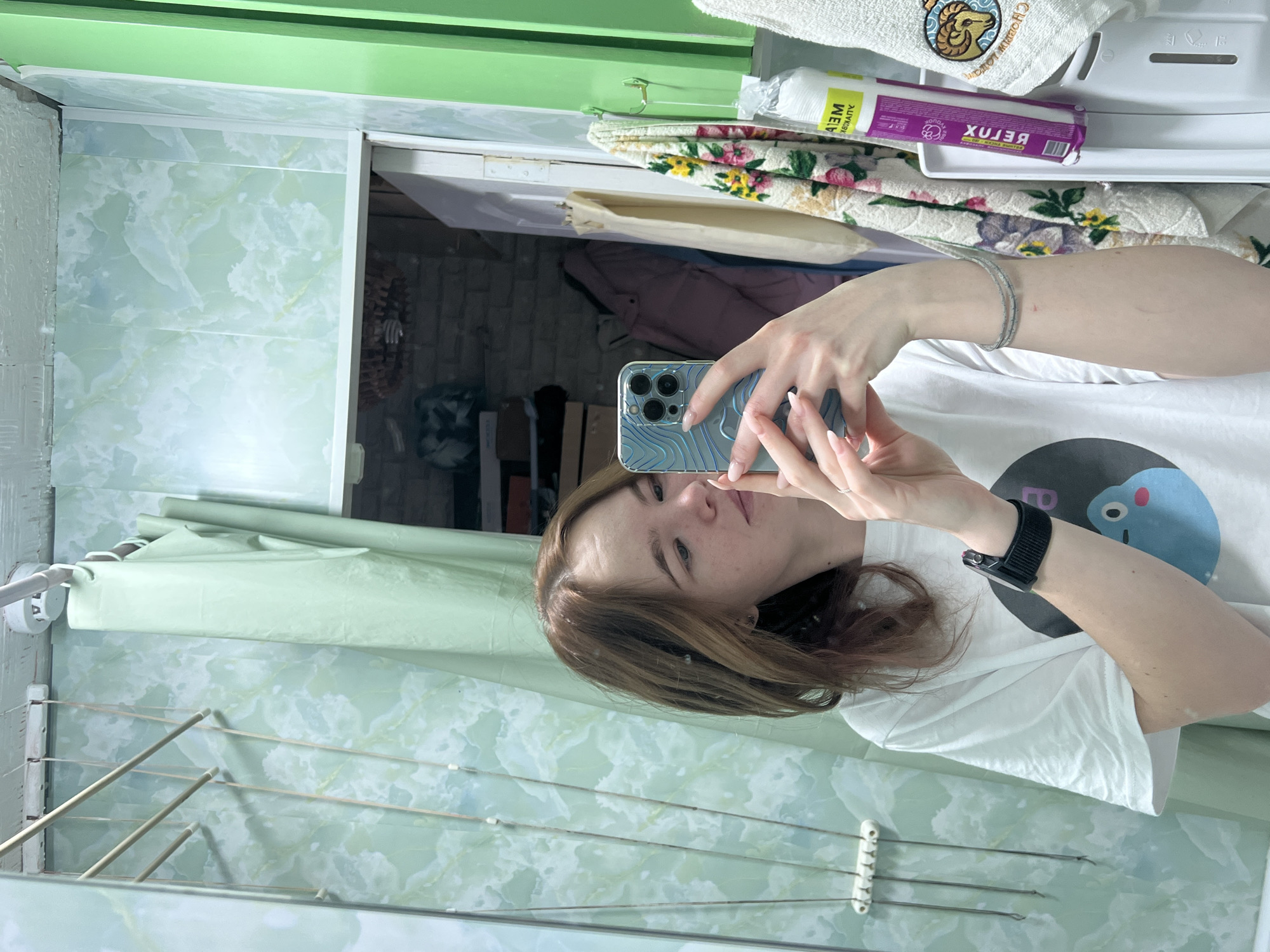 A person stands in a bathroom in what is presumably a residential building. The person holds a smartphone in front of them, likely taking a selfie. The bathroom walls are covered in light green and white marble-patterned tiles. A clothes drying rack is mounted on the wall to the left. A cabinet with towels and bathroom supplies is located above the person. Without external identifiers, it is not possible to independently confirm that this location is specifically within the city of Chaikovsky, Russia.