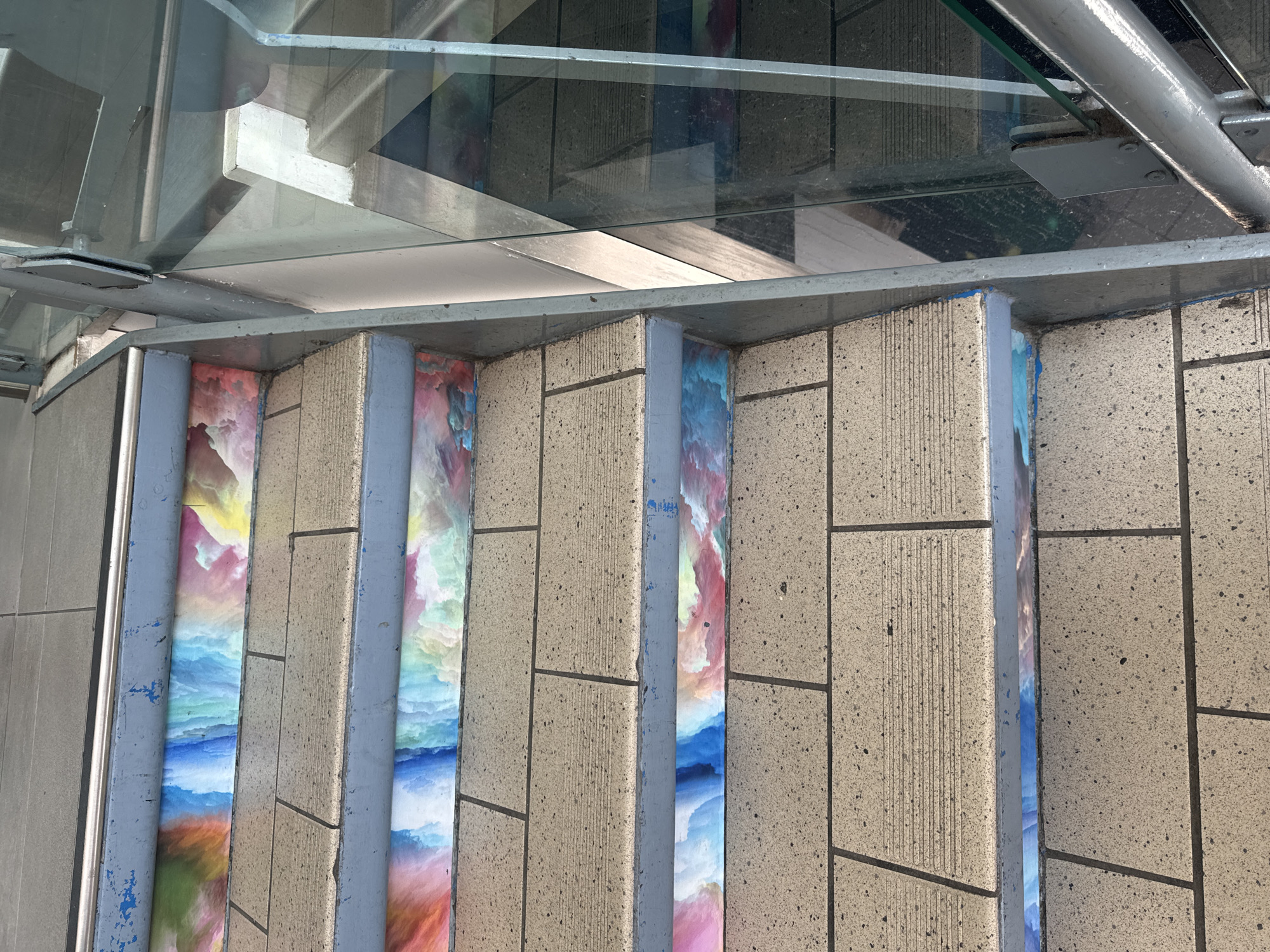 The image depicts a section of outdoor stairs with colorful, abstract artwork applied to the risers of each step. The steps themselves are made of light-colored, textured stone or concrete blocks laid in a running bond pattern. The risers feature vibrant swirls of blues, pinks, yellows, and whites, resembling a watercolor or cloud-like effect. A metal handrail runs along the left side of the stairs, painted a faded blue. To the right, a glass railing is visible, reflecting the stairs and the surrounding environment. The scene is likely located in an urban setting, possibly a public walkway or an entrance to a building. This image was captured on the 3rd of July, 2023, at approximately 11:00 AM in Waterford, Ireland.