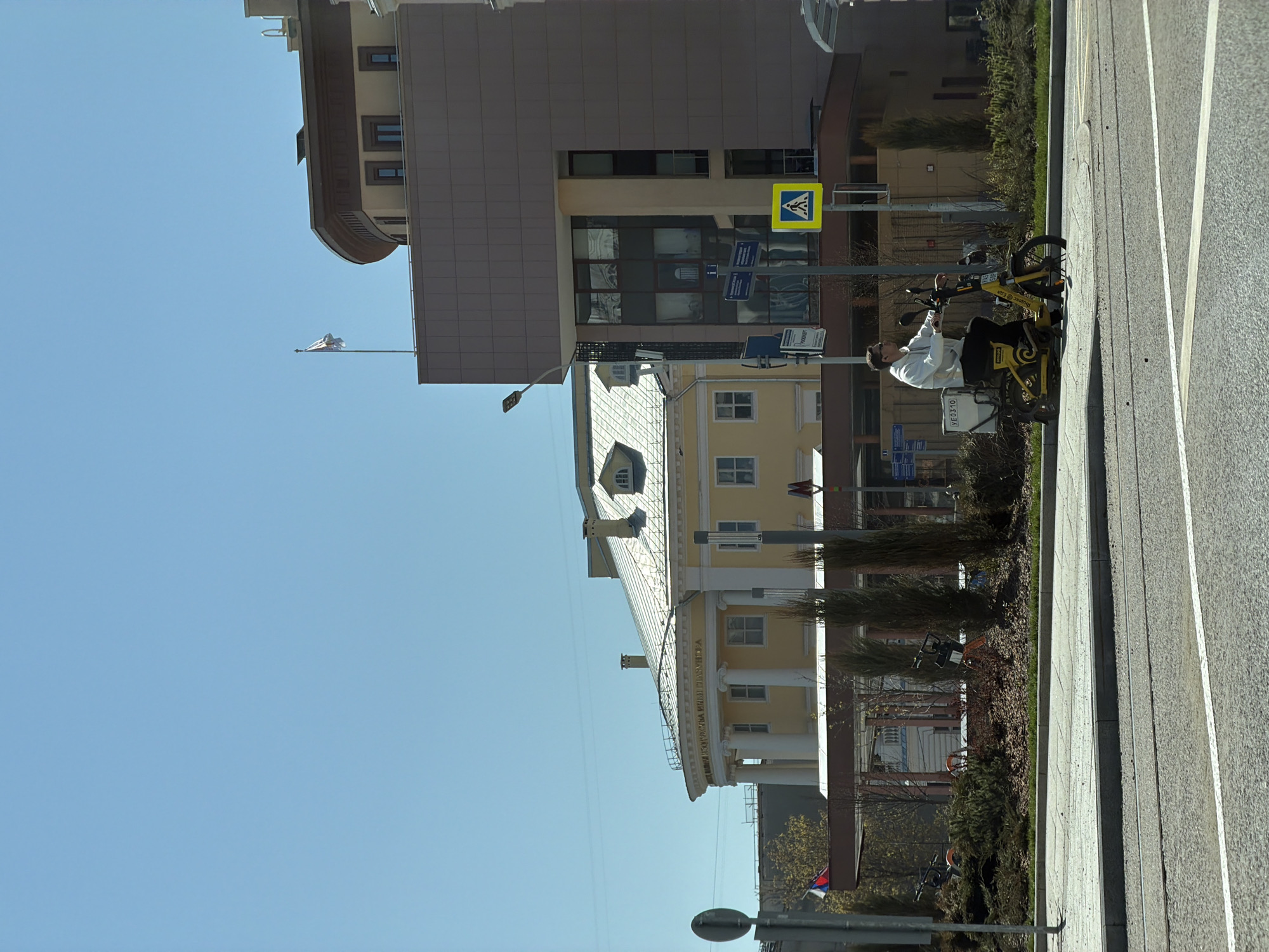 The image shows a street scene in Moscow, Russia, on a clear, sunny day. A man in a white jacket is seated on a yellow electric scooter on the sidewalk, which is located next to a multi-lane road. A pedestrian crossing sign is visible nearby. Behind the scooter, there are buildings, including a tan-colored building with a darker brown facade and a yellow building with white columns and architectural details. A flag is visible on a pole near the buildings. The sidewalk is concrete and the road is asphalt with painted white lane markers.