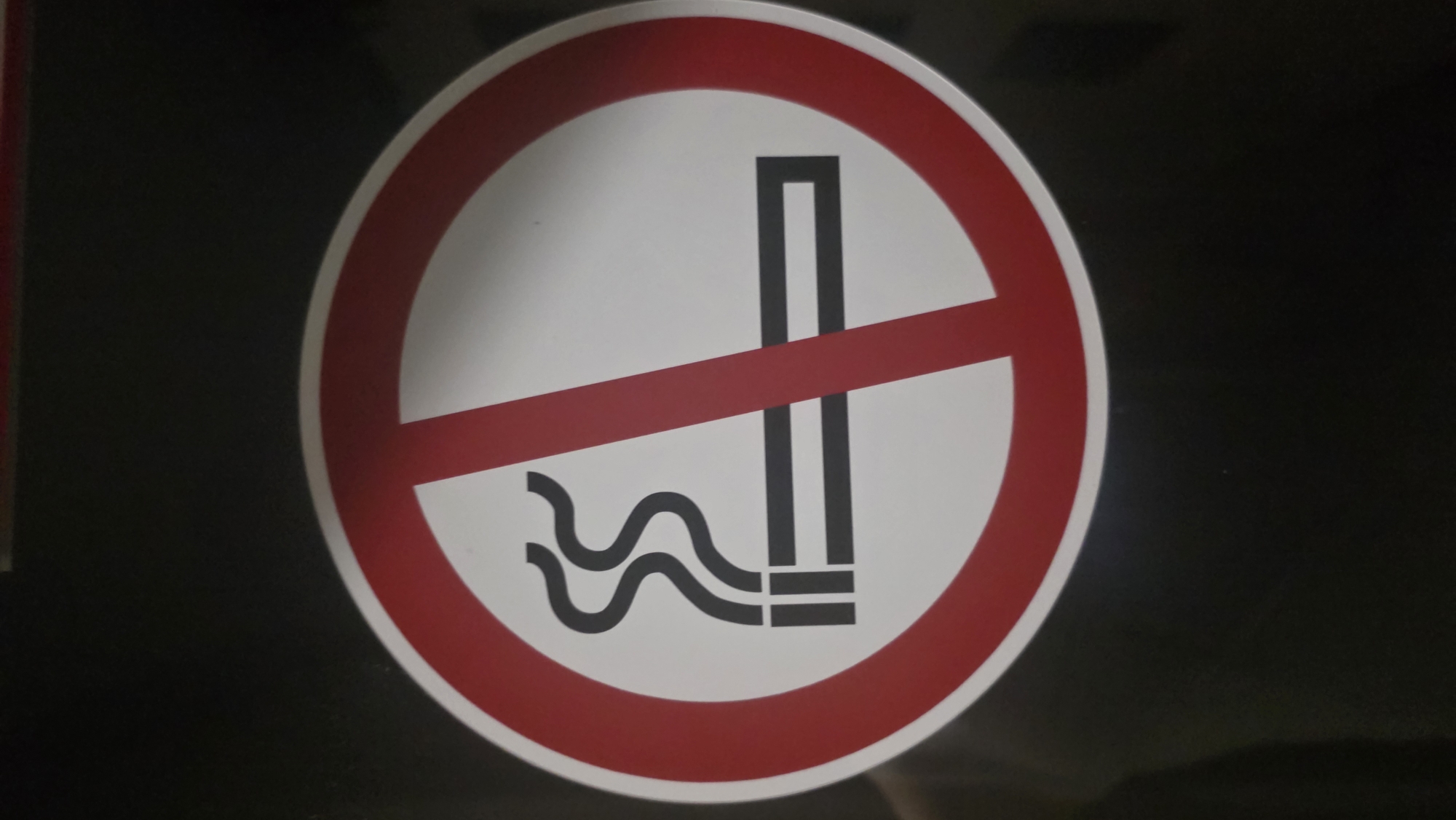 A no-smoking sign is displayed on a black background. The sign is circular with a red border and a white interior. A red diagonal line crosses the circle from the top left to the bottom right, indicating prohibition. Inside the circle, a black symbol depicts a lit cigarette with smoke rising from it. The cigarette is shown horizontally, with two horizontal lines representing the filter and body of the cigarette, and two wavy lines representing smoke. The sign is centrally located and appears to be a sticker or decal. There are no people or other objects visible in the frame. The lighting is even, and the image is in focus.