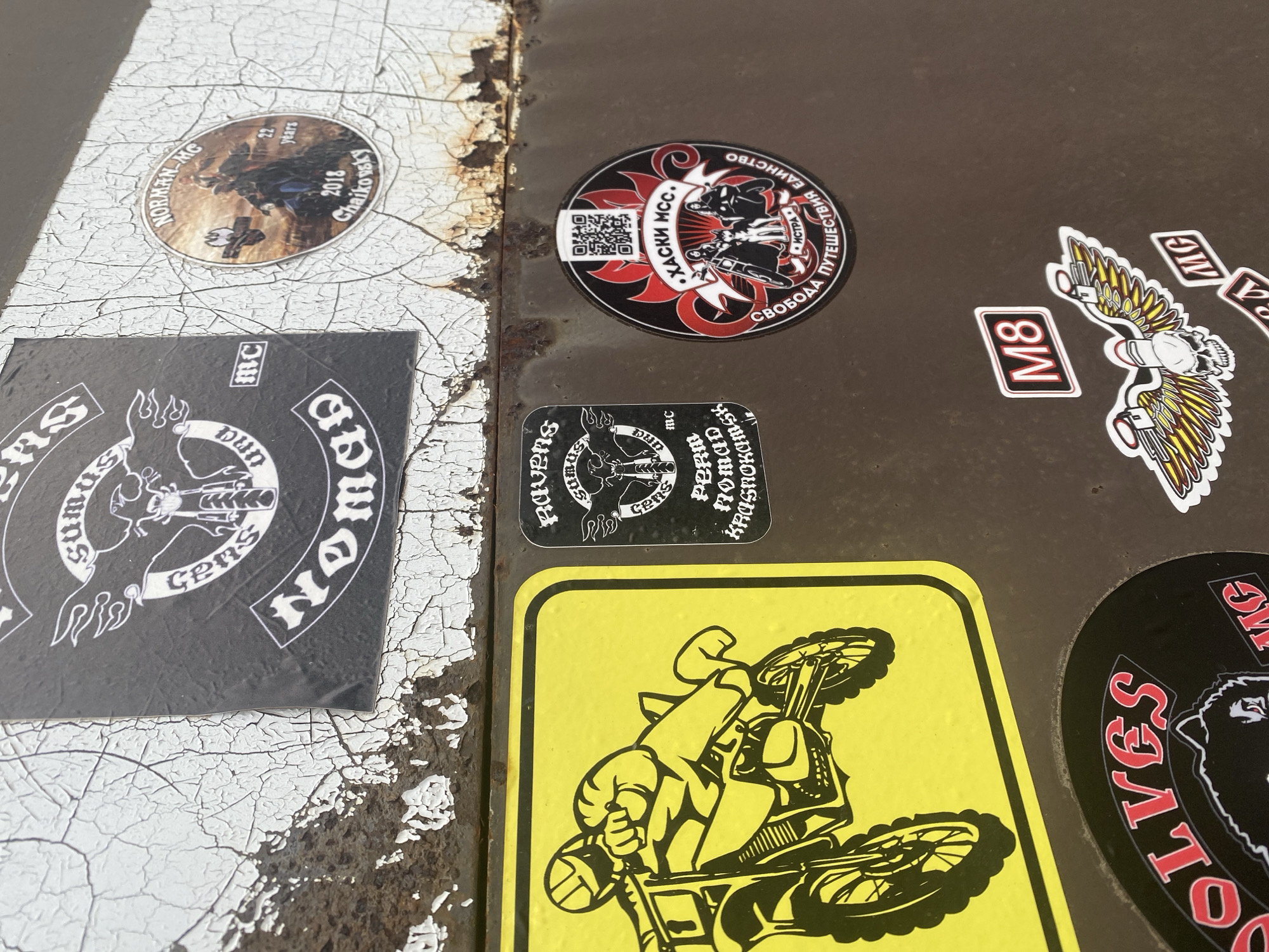 This image shows a close-up of a surface covered in stickers, likely belonging to or associated with motorcycle clubs (MC). The surface is divided into two sections, one painted white with a cracked texture and the other a darker, presumably metal, surface.
The stickers vary in size, shape, and design. Some are circular, others square or rectangular. There are stickers with logos of motorcycle clubs ("NOMAD Cens," "ХАСКИ МСС," "Norman-MG," "Wolves MG") and others with imagery related to motorcycles, like a rider on a dirt bike. The sticker for Norman-MG specifies "Chaikovsky" and the years 2018 and "22 Years," implying it is associated with the city of Chaikovsky, Russia. Some stickers contain text in Russian ("СВОБОДА ПУТЕШЕСТВИЯ ЕДИНСТВО"). The M8 and MG stickers feature images of skull with wings.
