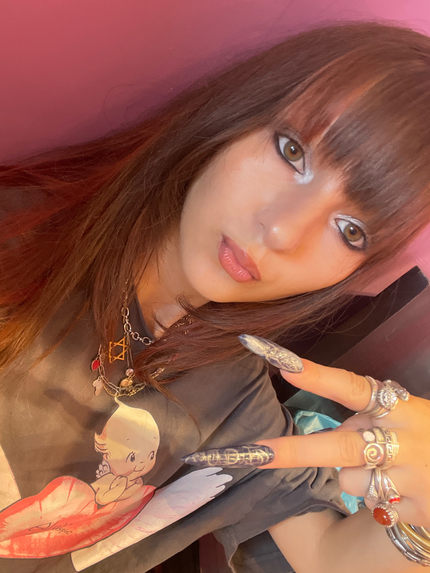 The image shows a close-up selfie of a young person with long, dark hair with reddish highlights, styled with bangs. They are wearing makeup, including silver eyeshadow and glossy lipstick. Several rings adorn their fingers, and a necklace with various charms is visible. The person is wearing a dark t-shirt with a graphic print of a cherubic figure lying on lips. The background appears to be a pink wall. The individual's left hand is raised, showing off their decorated fingernails and rings. The image does not contain any information that would allow for determination of its location in Raanana, Israel.