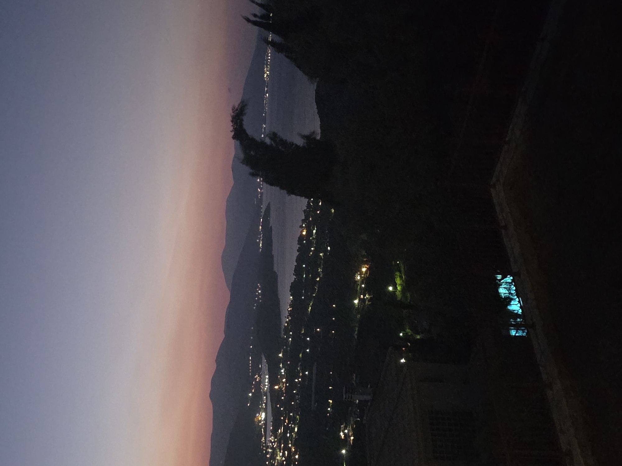 A wide shot of the Municipal Unit of Asini, Greece, is captured during dusk. The sky transitions from a pale blue at the top to a soft pink and orange hue closer to the horizon. In the distance, a body of water is visible, with rolling hills and mountains forming the backdrop. Along the shoreline and on the slopes of the hills, numerous lights from buildings and streetlamps illuminate the landscape, suggesting a populated area. In the foreground, dark silhouettes of trees and vegetation obscure parts of the view. On the left side of the foreground, a portion of a building with a tiled roof and a window with visible bars is seen. To the right of this building, a brightly lit turquoise pool or water feature is visible. The overall impression is a serene, twilight view of a coastal town.