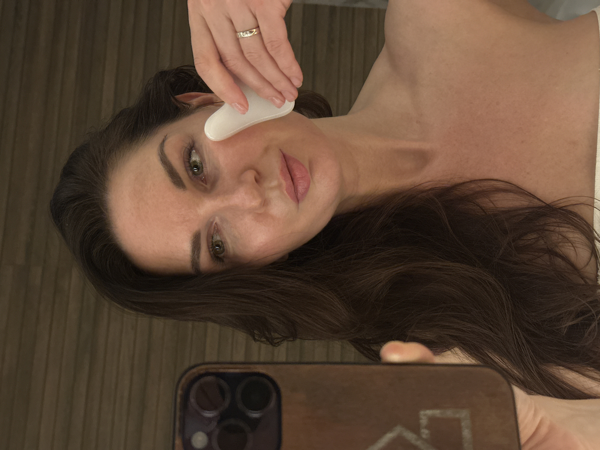 A woman is taking a selfie while holding a white gua sha tool to her face. She has dark brown hair, fair skin, and light brown eyes. She is wearing a gold ring on her left ring finger. The background is blurred and appears to be a wall with vertical lines. A portion of the phone being used for the selfie is visible in the bottom of the frame. There is no information in the image indicating its location is in Chaikovsky, Russia.