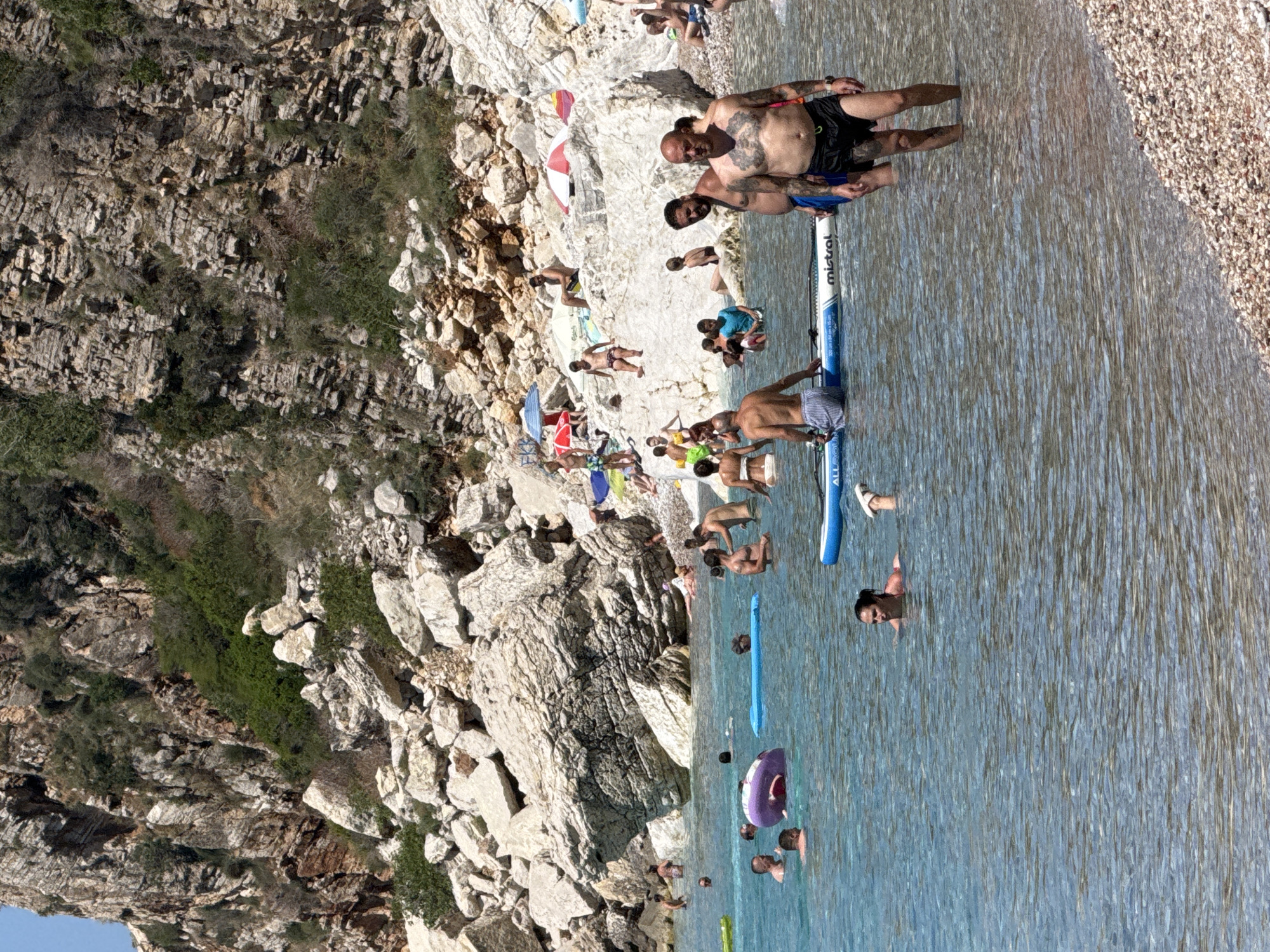 The image captures a crowded beach scene in Mirište, Montenegro, on a sunny day. The water is a clear turquoise, with people swimming, wading, and relaxing on inflatable floats. A paddleboard is visible in the foreground. The beach is composed of pebbles and large rocks, with a steep, rocky cliff face dotted with vegetation rising behind it. Numerous individuals are scattered across the water and along the shore, engaging in various leisure activities. Some are sunbathing on the rocks, while others are enjoying the water. Umbrellas and beach chairs are present on the shore, indicating a well-established recreational area. The overall atmosphere is one of a popular summer destination with a vibrant beach-going crowd.