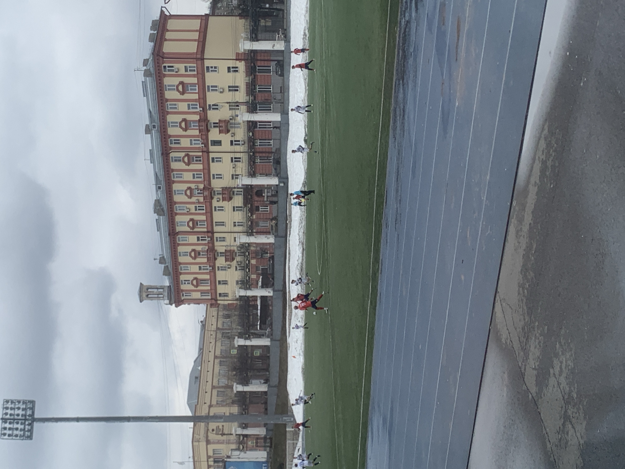 A medium shot shows a soccer match on a green field with players in various colored jerseys. The field is bordered by a running track and a snow patch near the base of a large building complex. In the background are apartment buildings in Izhevsk, Russia. The day appears overcast, and a large metal stadium light pole is visible in the foreground.