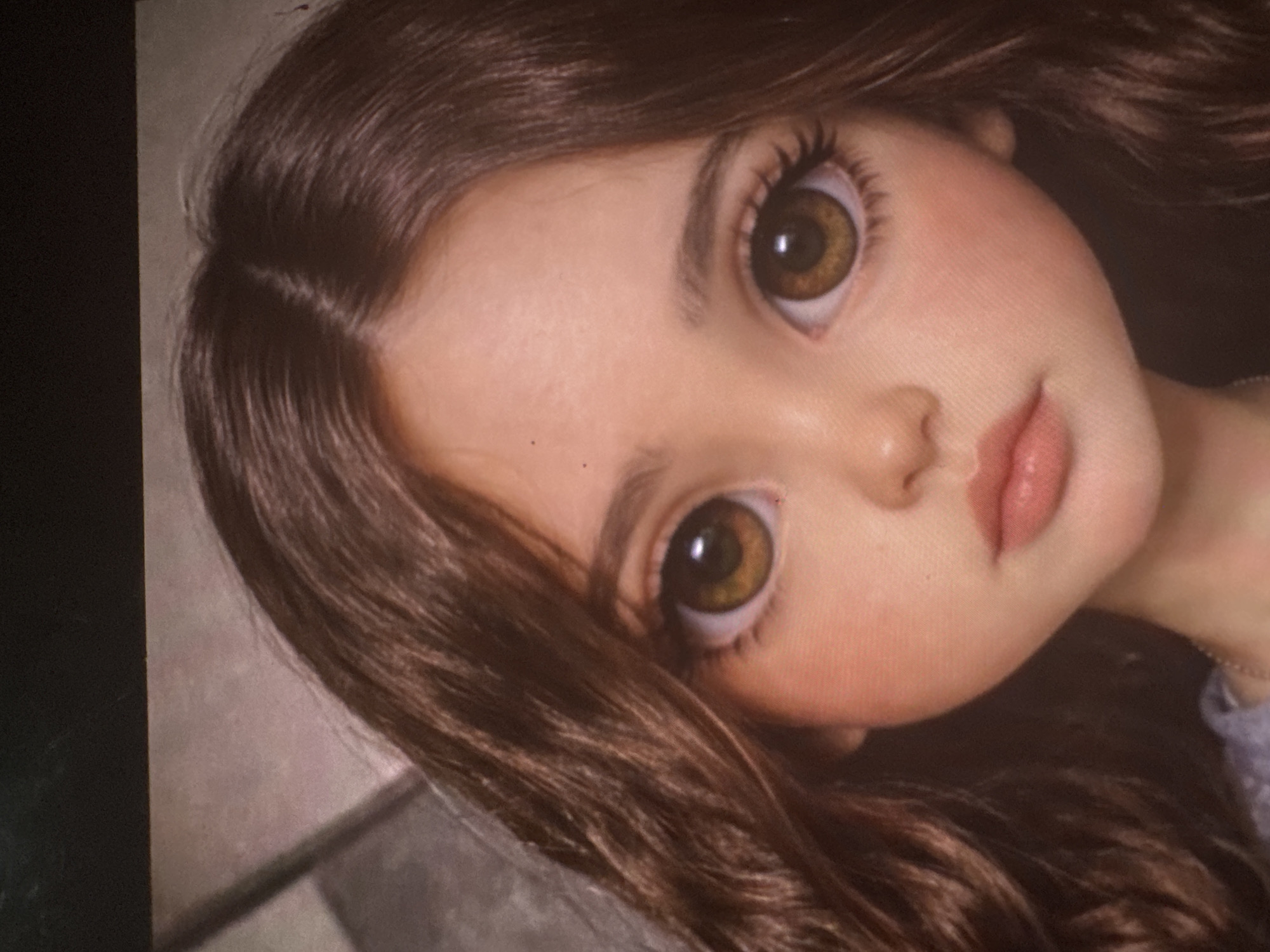 A close-up, diagonal shot features a highly detailed doll's face and upper chest. The doll has large, expressive brown eyes with prominent pupils and irises, framed by long, dark eyelashes and natural-looking eyebrows. Her skin tone is fair with a subtle blush on the cheeks and a natural texture visible. Her lips are full and a soft, light pink color. A small, sculpted nose sits centrally on her face.
The doll's hair is long, wavy, and a rich dark brown color, styled with a side part and falling around her face and shoulders. The hair strands appear individually rendered, adding to the realism. On the far right, a hint of a light-colored garment, possibly lavender or blue, is visible around the neck and shoulder area.
The background is indistinct and blurred, appearing as a light grey or beige tone on the left side and darker, muted tones on the right. The image itself appears to be a photograph taken of a digital screen, as subtle screen pixel patterns are discernible upon close inspection, particularly in the provided crops. The overall impression is one of innocence and serenity due to the doll's neutral expression and wide, gazing eyes. There is no discernible activity taking place, nor any visible text or specific location cues beyond an indoor setting.