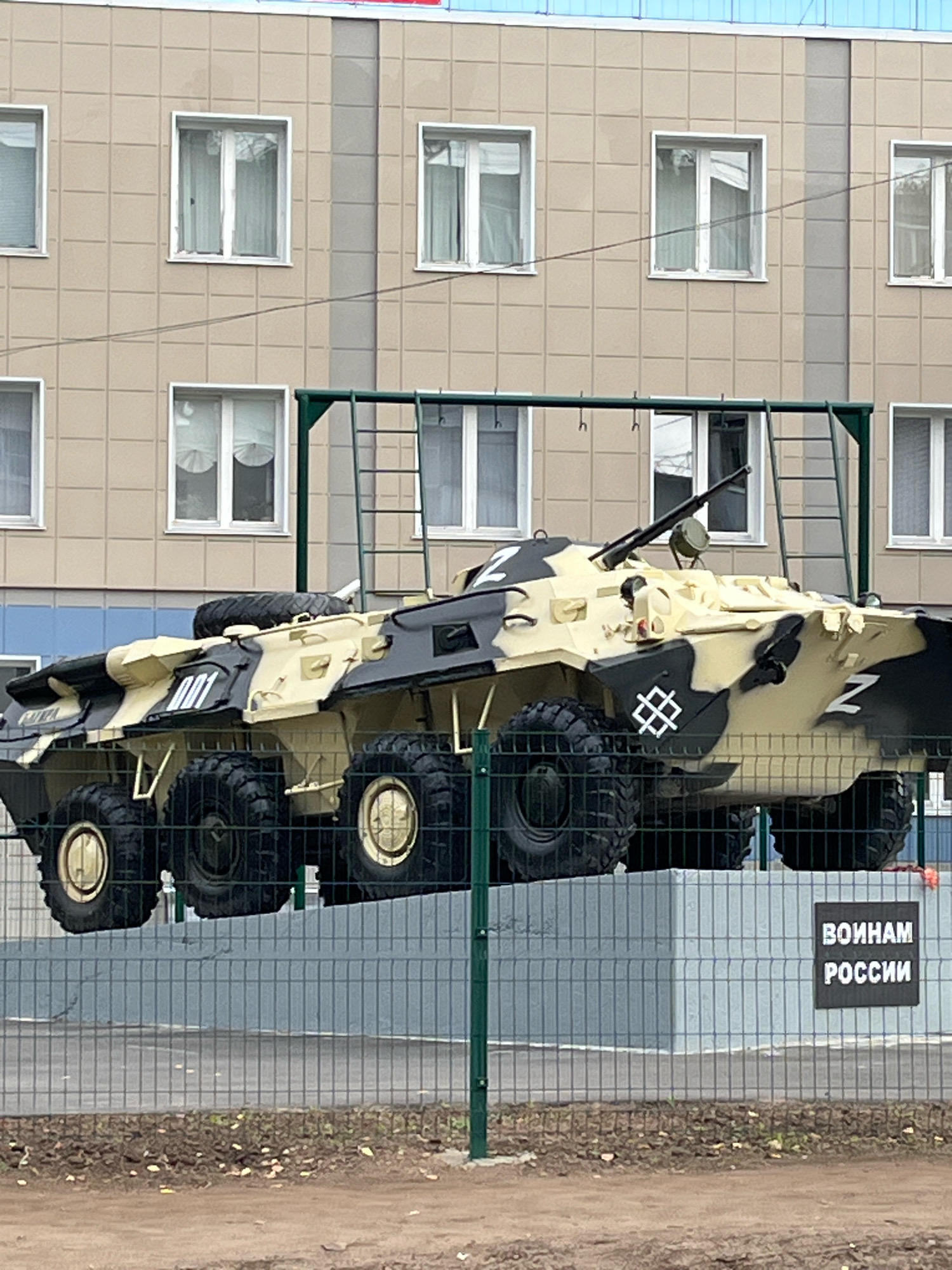A camouflaged military vehicle, possibly an armored personnel carrier, is displayed outdoors. The vehicle is painted in a pattern of tan, black, and gray. It has eight large tires and a mounted weapon system. On the side of the vehicle, there are white markings, including the letter "Z" and the numbers "001". There is also a white symbol that resembles a stylized square with interwoven lines. The vehicle is elevated and supported by a metal structure. In the background, there is a light-colored building with multiple windows. A dark sign with white text in Russian, reading "ВОИНАМ РОССИИ" (To the Warriors of Russia), is visible on a fence in front of a concrete structure. The scene appears to be in a public area, possibly a memorial or exhibition, located in Chaikovsky, Russia.