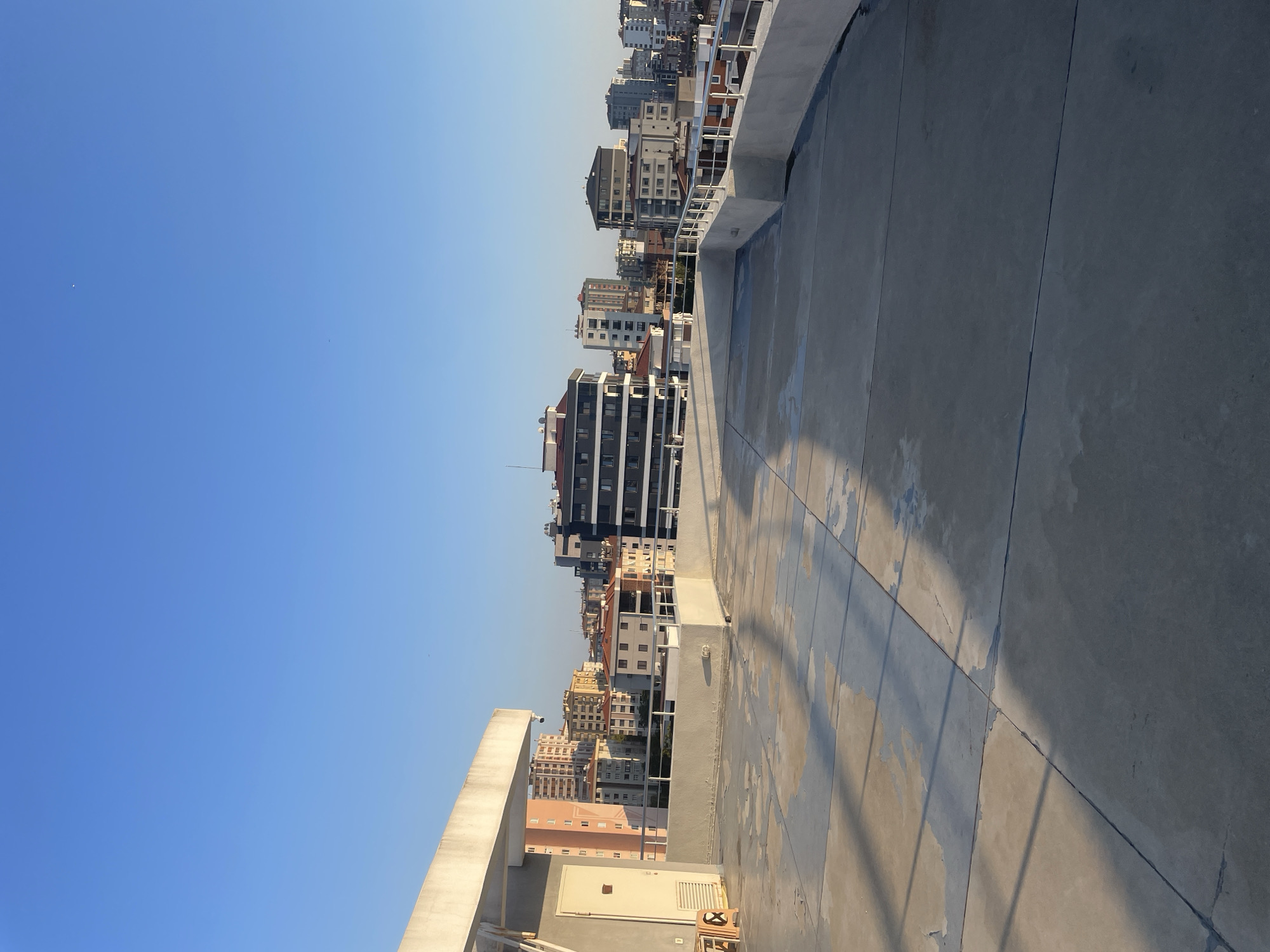 A low-angle, medium shot captures a rooftop terrace overlooking a dense urban cityscape under a clear blue sky. The terrace floor is made of large concrete slabs, some of which are stained and worn, with distinct lines indicating the separation between the slabs. Long shadows from an unseen structure stretch across the terrace, creating diagonal patterns. A white concrete railing runs along the edge of the terrace, with metal poles interspersed. On the left side of the terrace, a white door with a metal handle is set into a light-colored wall. To the left of the door, two orange plastic chairs are stacked. Beyond the terrace railing, a collection of multi-story buildings with various architectural styles and colors populate the background. The buildings range from modern, dark-colored structures to older, pastel-colored apartment buildings. The sky is a vibrant, cloudless blue, suggesting fair weather. The scene appears to be in Kadıköy, Turkey, a bustling district on the Asian side of Istanbul.