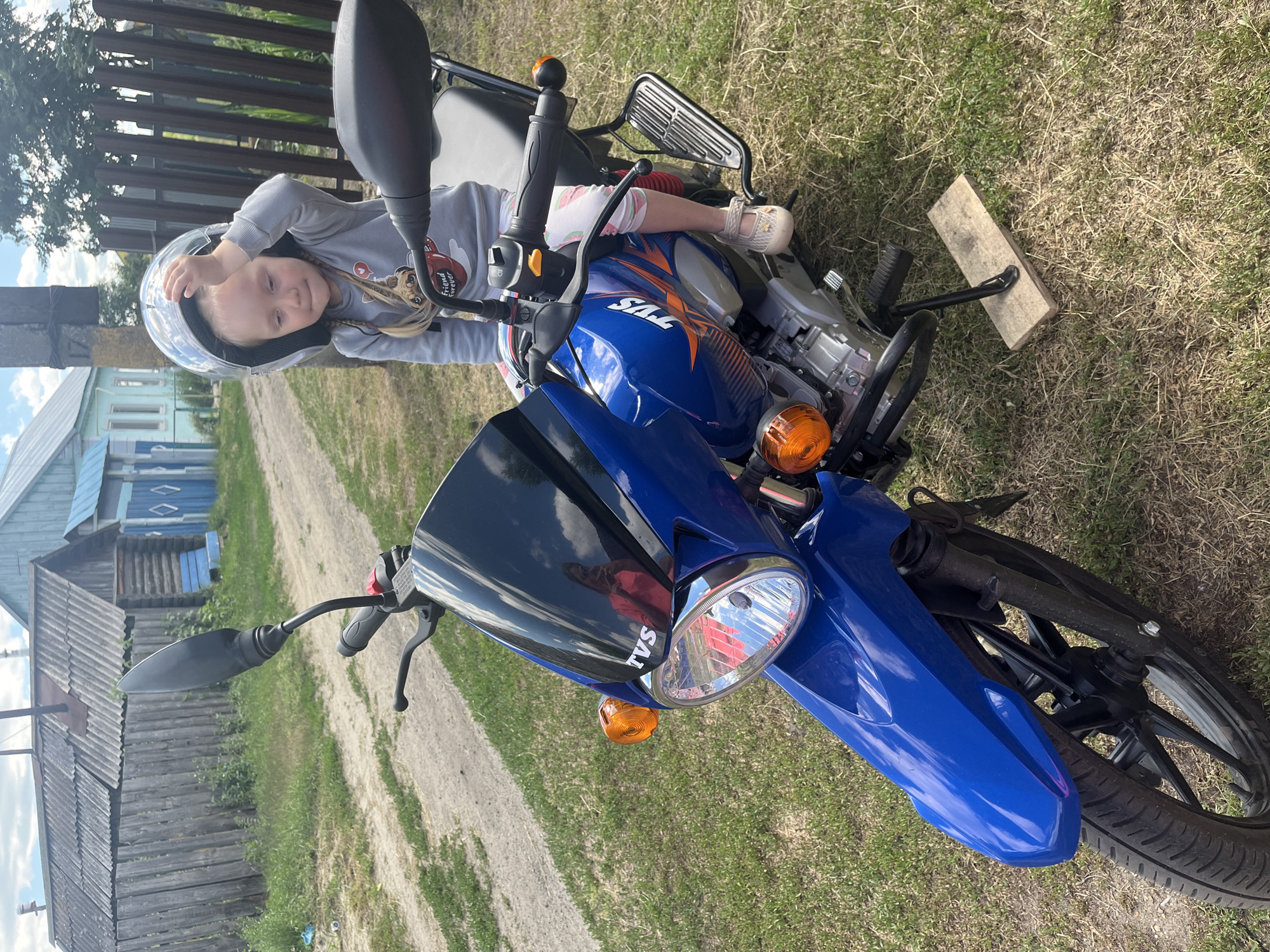 A young girl wearing a grey hoodie, white and pink patterned pants, and a clear helmet is sitting on a blue TVS motorcycle. She is leaning back, with her right arm resting on the handlebar and her legs extended forward. Her hair is in braids, and she appears to be smiling, looking towards the camera. The motorcycle is parked on a grassy area next to a dirt path, with a wooden fence and houses in the background. A wooden plank is placed under the motorcycle's kickstand. The scene appears to be outdoors during the daytime, with sunlight casting shadows. The text "TVS" is visible on the side of the motorcycle. The location is identified as Сухринское, Russia.