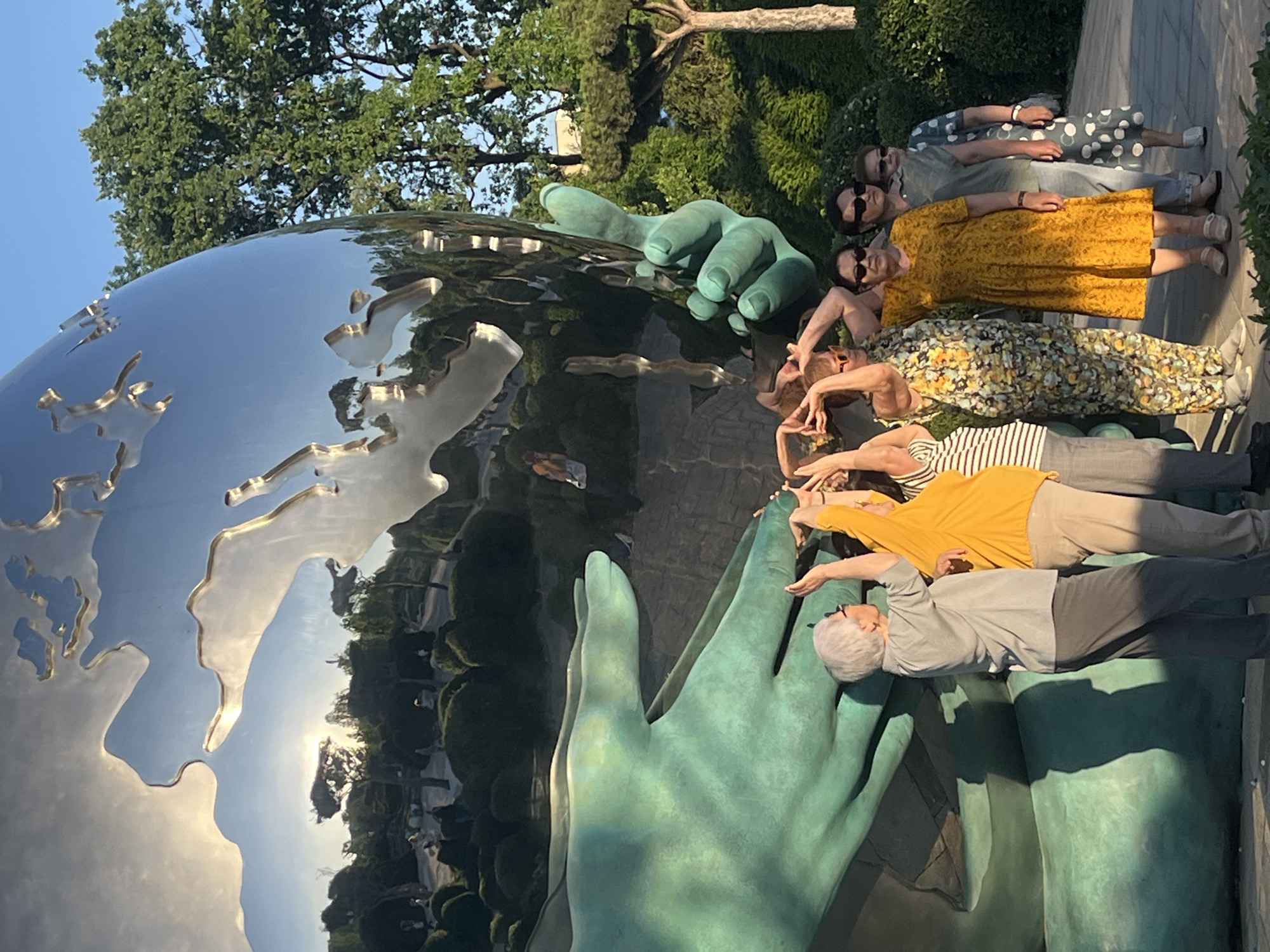 The image captures a group of six women posing alongside a large, distinctive outdoor sculpture under bright sunlight. The prominent sculpture features a highly reflective, metallic globe with etched landmasses, supported by two enormous, weathered teal-colored hands. The globe's polished surface reflects the clear blue sky, surrounding trees, and distorted figures of the people and environment.
The women, ranging from middle-aged to elderly, are arranged in a line to the right of the sculpture. They are dressed in various casual, summery outfits, including patterned dresses and tops, and several are wearing sunglasses. Most of them are actively interacting with the sculpture, extending their arms towards or resting them on the large teal hands, seemingly mimicking a gesture of support or connection with the globe. Their postures suggest they are posing for a photograph, with some looking towards the camera and others engaged with the sculpture.
The scene is set outdoors in what appears to be a park or garden, indicated by the lush green trees and foliage forming the background. The ground beneath the women seems to be a paved or wooden surface. The lighting suggests late afternoon or early evening, with strong, warm sunlight casting noticeable shadows from the individuals onto the ground. The weather is clear and sunny. Based on the provided context, this sculpture is located in Krasnodar, Russia. No visible text is present in the image.