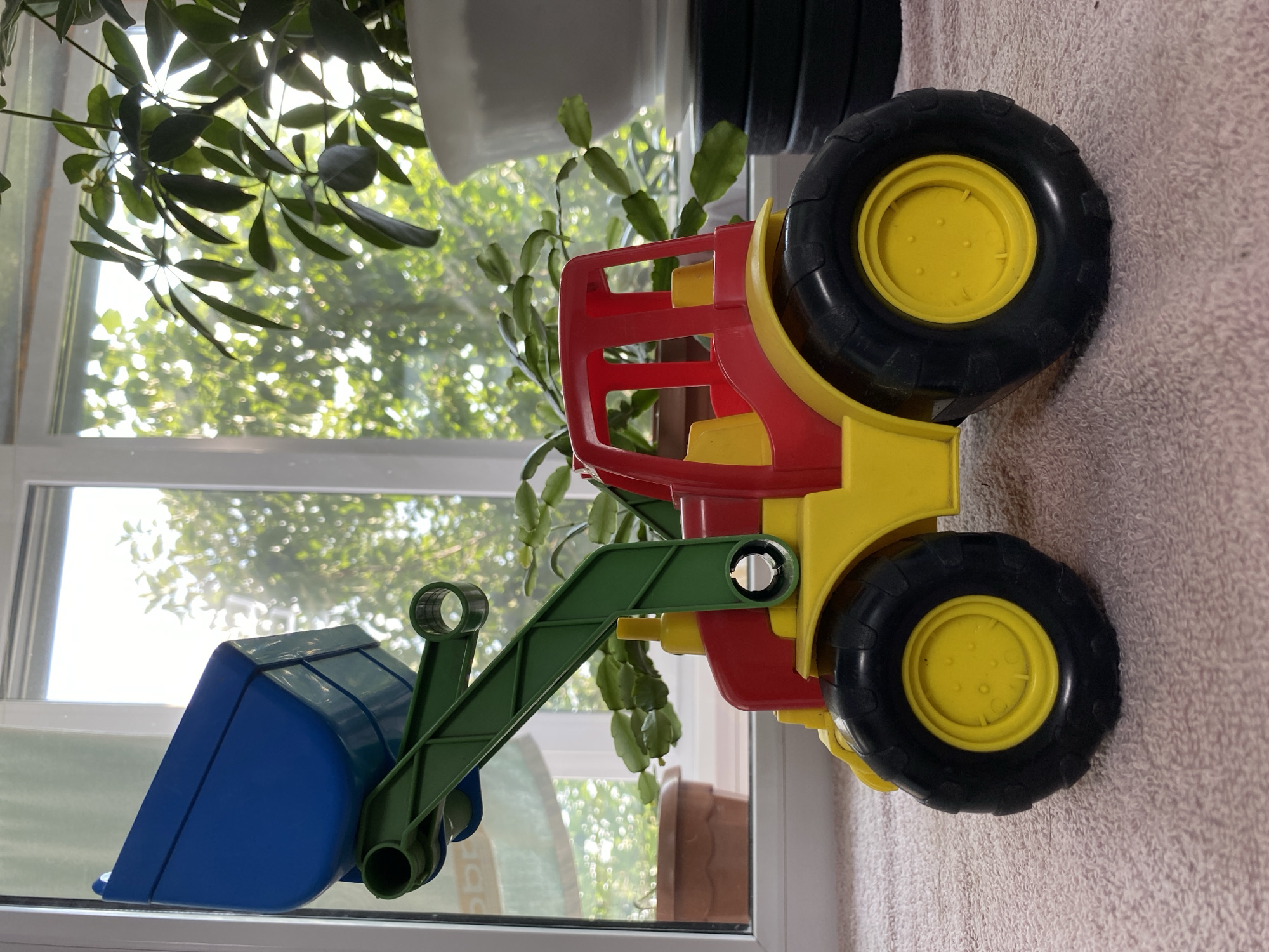 A bright red and yellow toy tractor with a blue bucket on a green arm sits on a pink carpet. The tractor has large black tires with yellow hubs. In the background, a window overlooks a sunny outdoor scene with green trees and foliage. There are also houseplants visible, one in a large white pot and another in a smaller brown pot. The scene is well-lit by natural light streaming through the window. There is no indication of the specific location in Бескөл ауылдық округі, Kazakhstan.