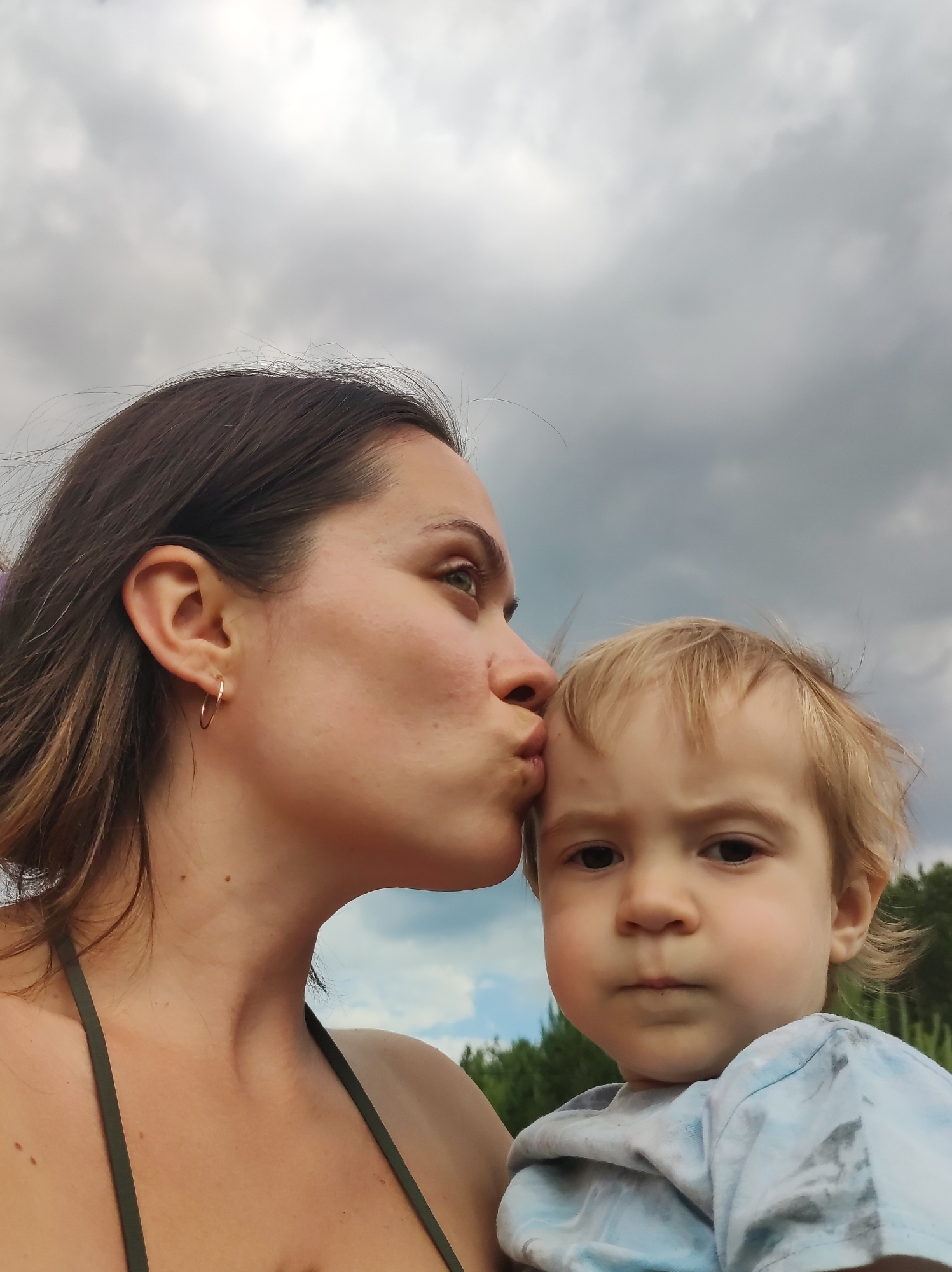 In this media file, a woman is shown kissing the forehead of a young child. The woman has dark brown hair and is wearing a green bikini top. The child has blonde hair and is wearing a light blue tie-dye t-shirt. Both individuals are looking towards the camera, though the child's expression is more direct and serious, while the woman's eyes are looking slightly off to the side.
The setting appears to be outdoors, with a cloudy sky in the background. Hints of green trees and foliage are visible behind the subjects, suggesting a natural or park-like environment. The lighting suggests it could be daytime, possibly late afternoon given the diffused light. The overall mood conveyed is one of affection and tenderness between the woman and child. There is no visible text in the image.