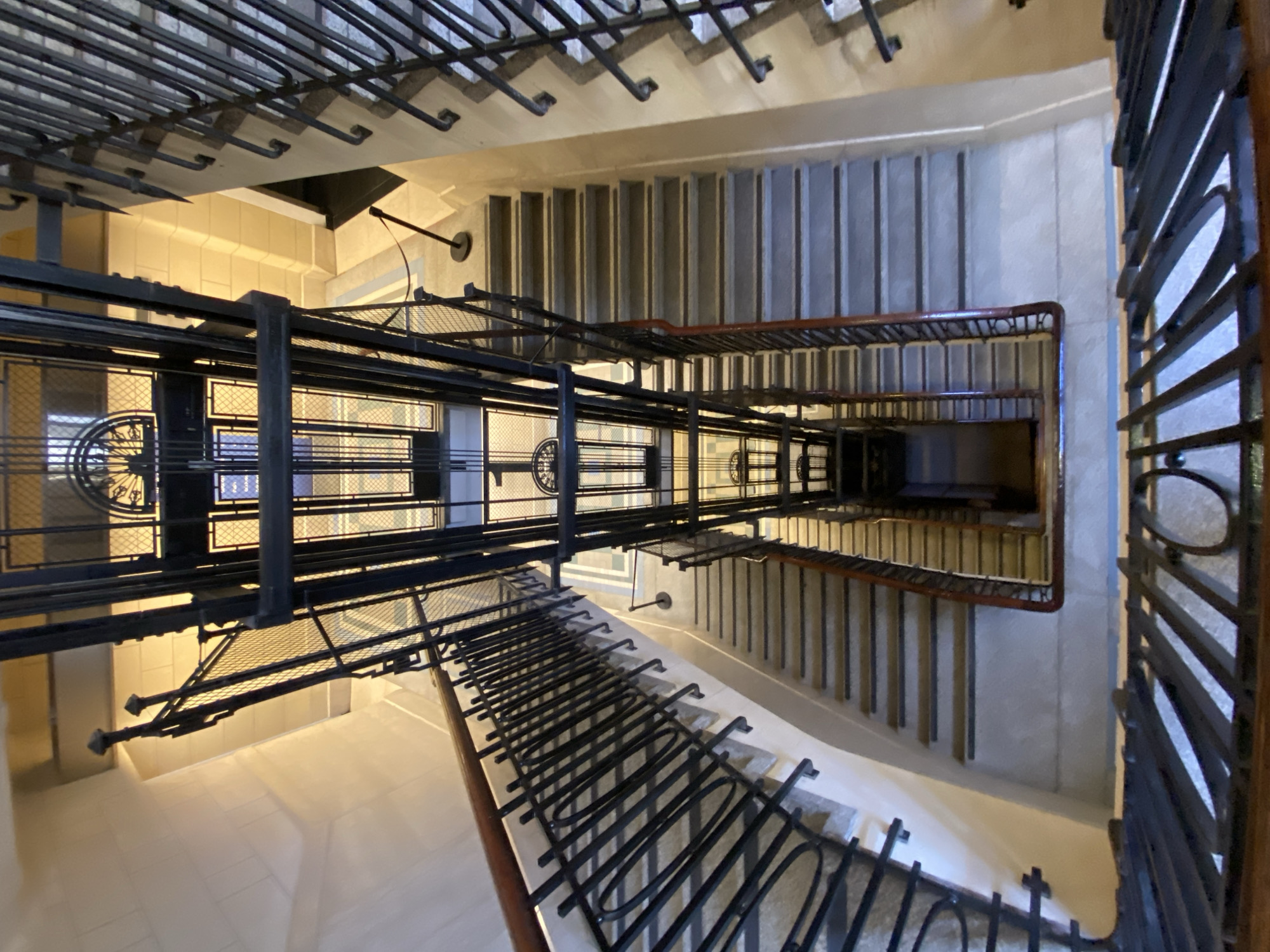 The image depicts the interior of a building in Buenos Aires, Argentina. It appears to be a multi-story building, possibly an apartment complex or office building. The viewpoint is from above, looking down into a central open area, possibly a stairwell or elevator shaft. The dominant architectural feature is a vintage-style elevator with a black metal cage. Staircases with metal railings are visible wrapping around the central space, leading down to lower floors. The walls are a neutral color, with some areas showing signs of age or wear. The floors appear to be tiled or stone. The lighting is somewhat diffused, suggesting natural light coming from windows or a skylight. The overall impression is of an older building with preserved architectural details. There are no people visible in the image.