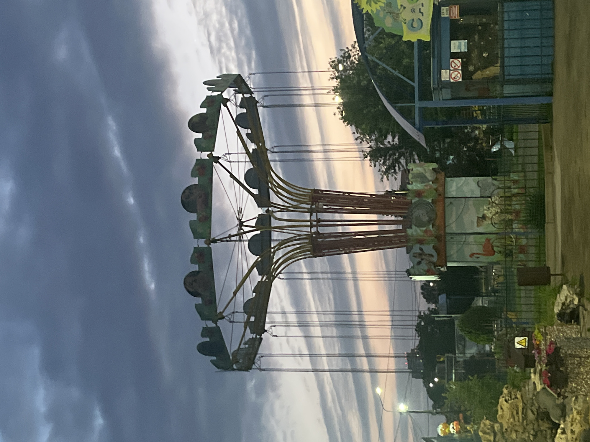 The image captures an amusement park swing ride in Krasnodar, Russia, during twilight. The sky displays a gradient of dark gray clouds at the top transitioning to lighter hues of pink and orange near the horizon, indicating the setting sun. The swing ride, painted in shades of green and yellow, dominates the center of the frame. Its circular seating area is suspended by numerous chains, which hang vertically against the sky. The central support structure is red and yellow. In the foreground, a low fence separates the amusement park from the viewer. Beyond the fence, trees and buildings are visible, including a blue kiosk with signage. There are no people clearly visible in the image, but the overall atmosphere suggests a quiet evening at the park.