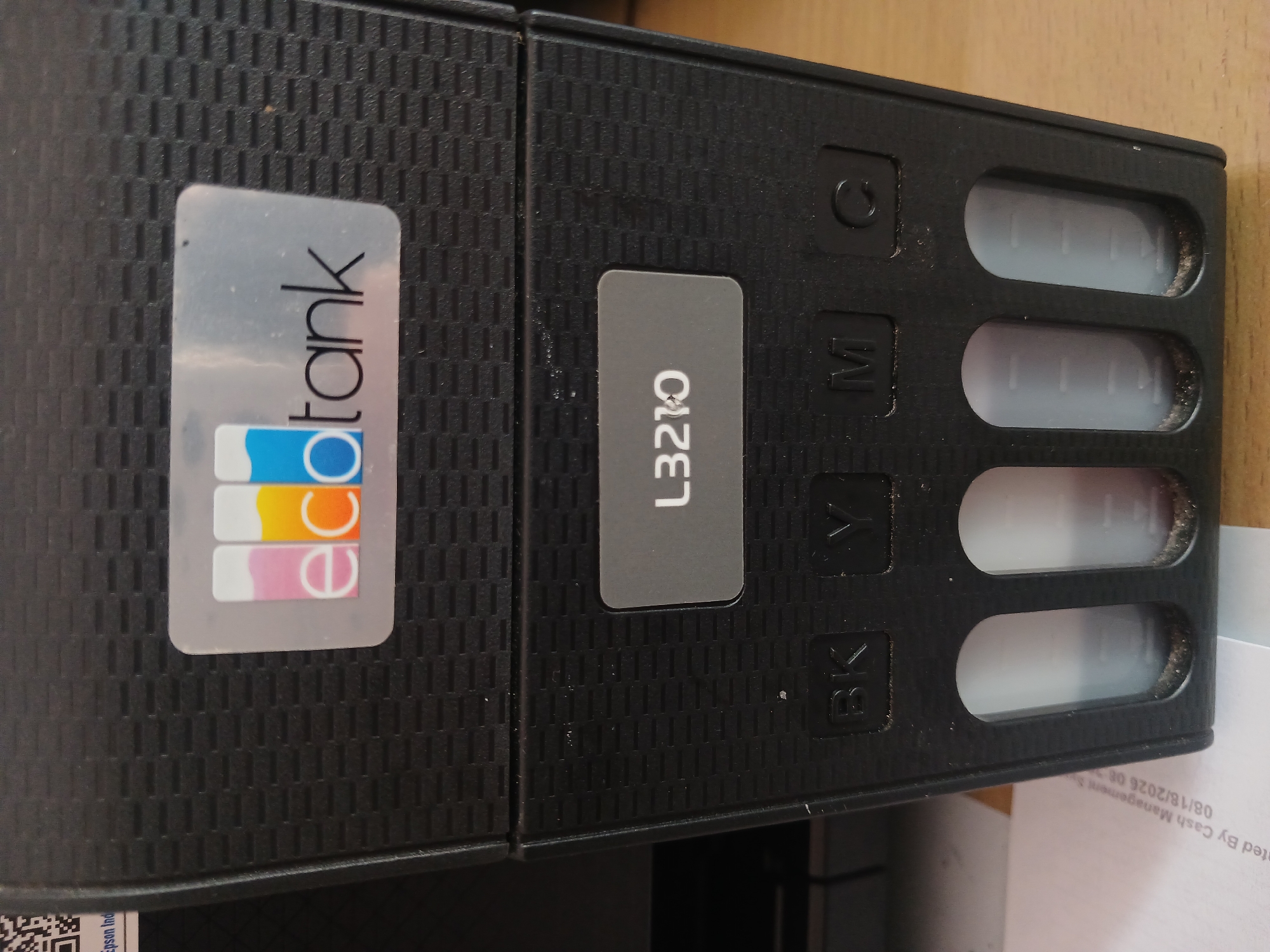 The image shows a close-up view of the front of a black Epson EcoTank printer. A label with the "EcoTank" logo, featuring colored ink bottles (pink, yellow, and blue), is visible on the upper section of the printer. Below this, a gray label displays the model number "L3210". Further down, there are four indicator slots labeled "BK", "Y", "M", and "C", corresponding to Black, Yellow, Magenta, and Cyan ink. Beneath these labels are the transparent ink tanks, showing the ink levels. The printer appears to be placed on a wooden desk, with a piece of paper partially visible on the left side. The overall environment suggests an indoor setting, likely an office or home. There are no people visible in the image. The location in Watugede, Indonesia is not evident from the image content.