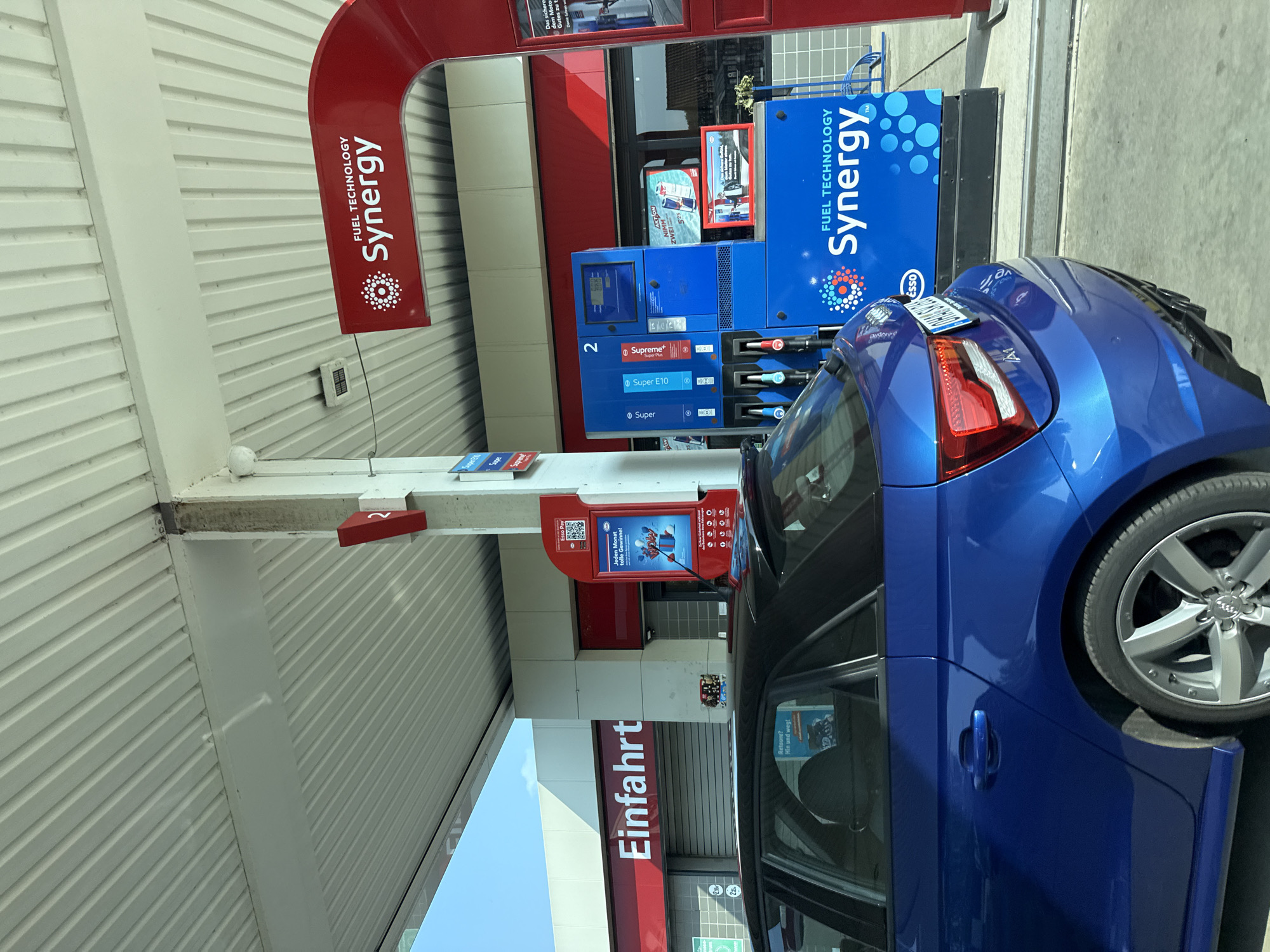 The image depicts a blue Audi A1 hatchback parked at an Esso gas station. The car is positioned in front of a blue fuel pump with "FUEL TECHNOLOGY Synergy" branding. The gas station canopy is visible overhead, featuring ribbed white panels and red accents. A sign with the word "Einfahrt" (entrance) is visible on a building in the background. Another sign on the pump displays a QR code and promotional text related to "Esso Pay" and a monthly prize draw. The scene is brightly lit by daylight, suggesting an outdoor setting. The car's license plate is partially obscured. The location is identified as a gas station in Bad Sachsa, Germany, specifically on the Harzburger Straße.