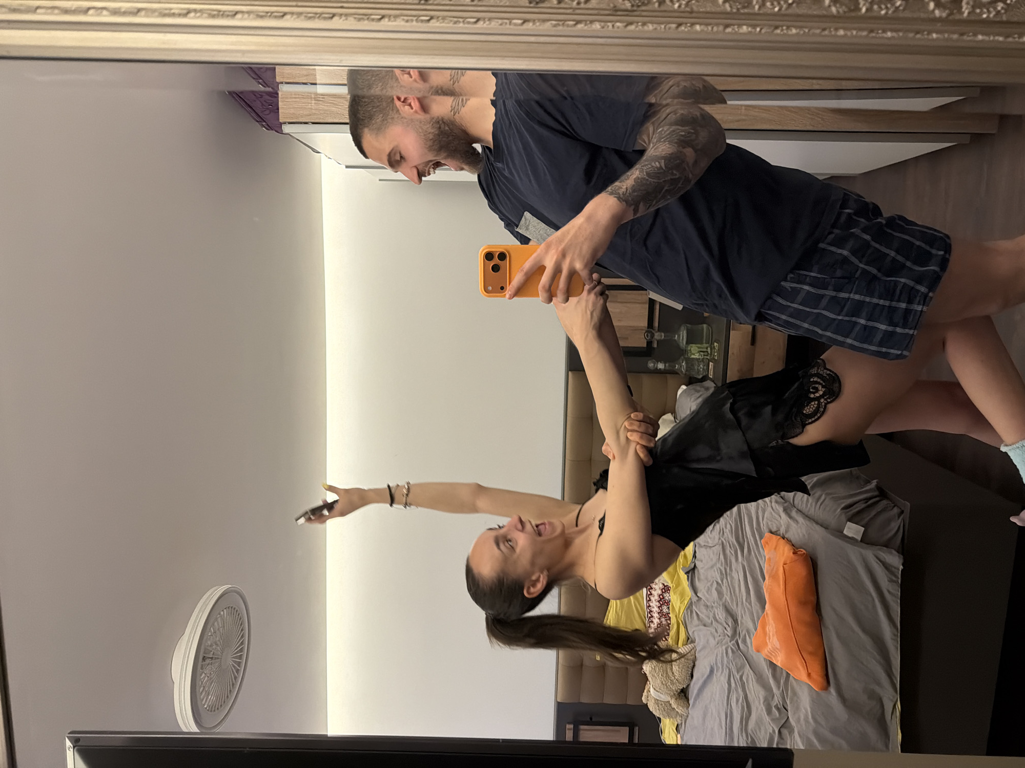 A mirror selfie captures a man holding a woman by the arms in what appears to be a bedroom. The man, on the right side of the reflection, holds an orange phone and has tattoos on his left arm. He is wearing a navy t-shirt and plaid shorts. The woman is on the left, and seems to be lifted into a pose. She wears a black outfit with lace details. A white ceiling fan is visible on the wall. The room has a bed with grey bedding and orange and yellow items. It is impossible to determine the exact location in Sofia, Bulgaria, from the provided image.