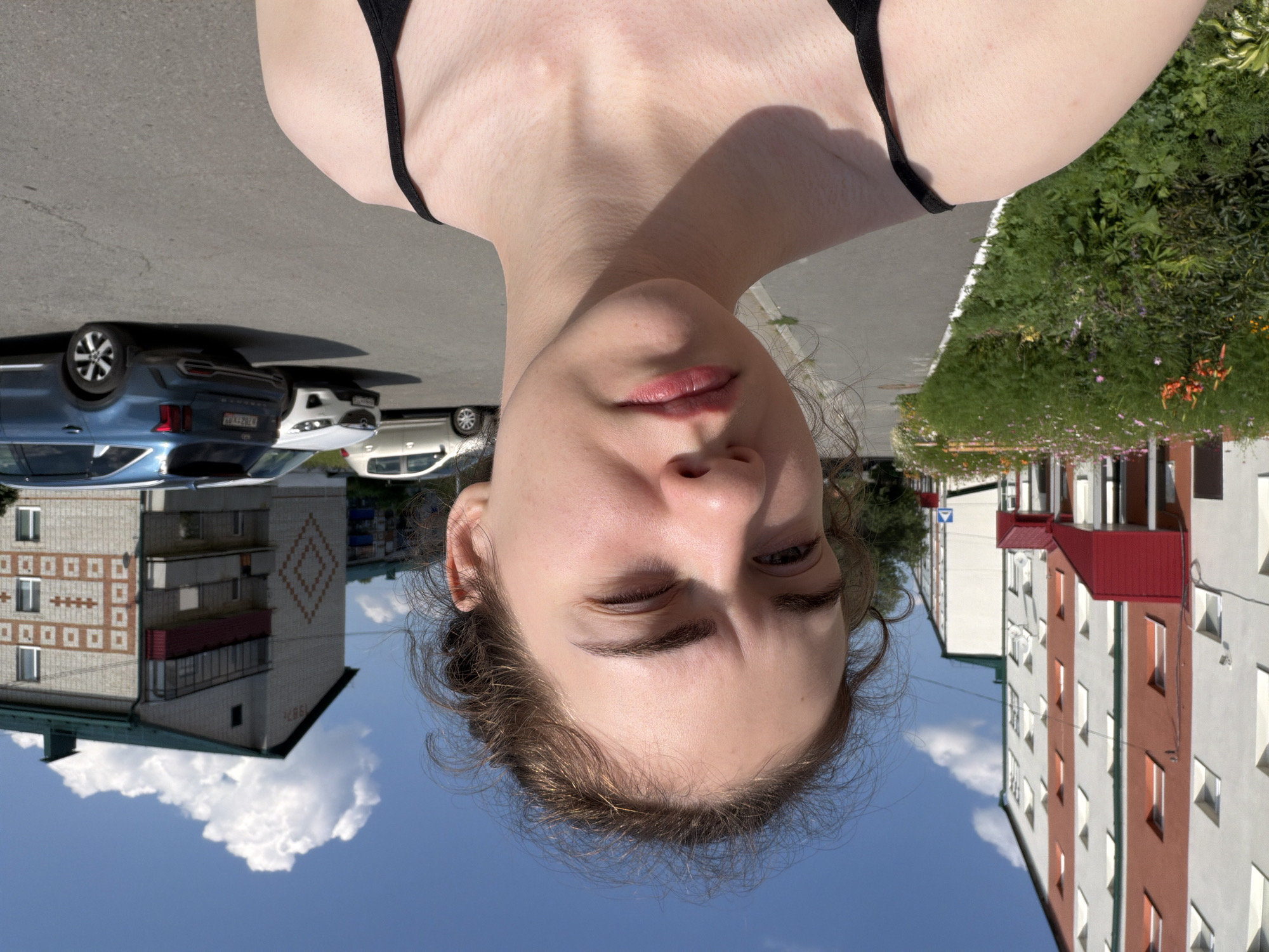 The image depicts a young woman taking a selfie outdoors on a sunny day. She is standing on a paved surface, likely a sidewalk or parking lot, with residential buildings visible in the background. To her left, a row of multi-story apartment buildings with white and red facades lines the street. A well-maintained flower bed with various colorful blooms runs alongside the sidewalk. To her right, several cars are parked, including a blue SUV and other passenger vehicles. The sky is clear with a few white clouds. The overall scene suggests an urban residential area. The image is believed to have been taken in Zavodoukovsk, Russia.