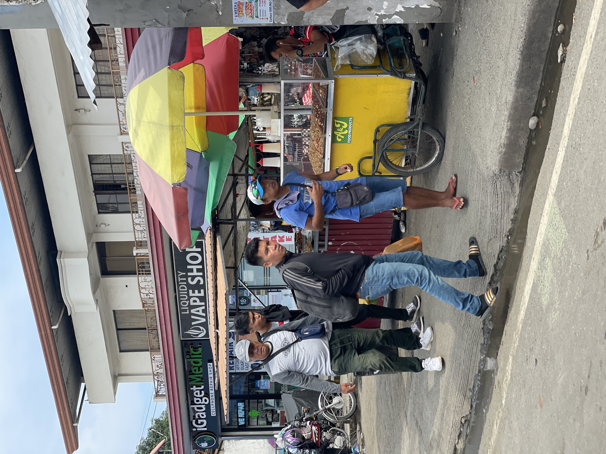 This detailed description outlines a street scene in Davao City, Philippines, featuring people, commercial establishments, and a vendor stall under an overcast sky. In the background, a two-story white building with a corrugated metal roof is visible. Its upper floor has multiple windows and a small balcony with an ornate railing. The ground floor houses two businesses: "iGadget Medic CELLPHONE REPAIR SHOP" (left, with a green and black sign featuring an Apple logo) and "LIQUIDITY VAPE SHOP" (right, with a black and white sign). The "iGadget Medic" storefront also displays "WE REPAIR" and "AFFORDABLE, GUARANTEED, FAST" signage. A small "18+" sticker is visible on the vape shop window. A brown awning extends below these shops. On the paved sidewalk, several individuals are present. From left to right: 1. A person wearing a purple helmet and dark clothing is crouched near a red and chrome motorcycle, partially visible on the far left. 2. An older man with a gray cap, gray and white long-sleeved shirt, and dark green cargo pants walks towards the right, carrying a dark blue sling bag. He wears white sneakers. 3. Behind him, a younger man with dark hair wears a black t-shirt over a white undershirt, dark pants, and black sneakers. 4. A young man with dark hair, wearing a black long-sleeved athletic jacket with three white stripes, blue jeans, and black Adidas slide sandals, walks towards the right. He carries a dark gray backpack on his left shoulder. 5. To the right, a man in a blue cap with sunglasses, a blue t-shirt, and blue denim shorts stands by a yellow food cart. He has a dark gray sling bag and a white towel over his left shoulder, holding a small white item in his left hand. He wears pink flip-flops. 6. Another man, wearing a black and red patterned t-shirt and dark pants, leans on the right side of the yellow food cart, appearing to be using white earbuds. The prominent yellow food cart, labeled "A&J FOOD PRODUCTS" in green, is a tricycle-style vendor cart. It features a clear glass display case on top, containing various fried or roasted food items. Red and white squeeze bottles are placed on the cart's surface. Multi-colored market umbrellas (yellow, red, green, purple, blue) are deployed above the food cart and extend further right, indicating additional stalls. A clear plastic bag containing items hangs from the right side