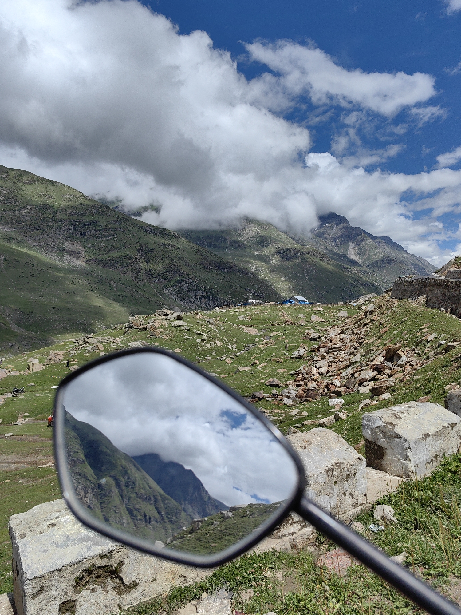The image, captured in Gramphu, India, presents a mountainous landscape under a partly cloudy sky. The foreground is dominated by the reflection in a motorcycle's rearview mirror, which shows lush green mountainsides and billowing white clouds. The actual scene outside the mirror features a rocky, green terrain with scattered boulders. In the mid-ground, several figures are visible, appearing to be on foot or riding motorcycles. A cluster of buildings, including a blue structure, is situated further in the distance. The mountains in the background are steep and covered in verdant vegetation, with some peaks partially obscured by clouds. A stone wall runs along a ridge on the right side of the image. The overall impression is that of a remote, scenic location.