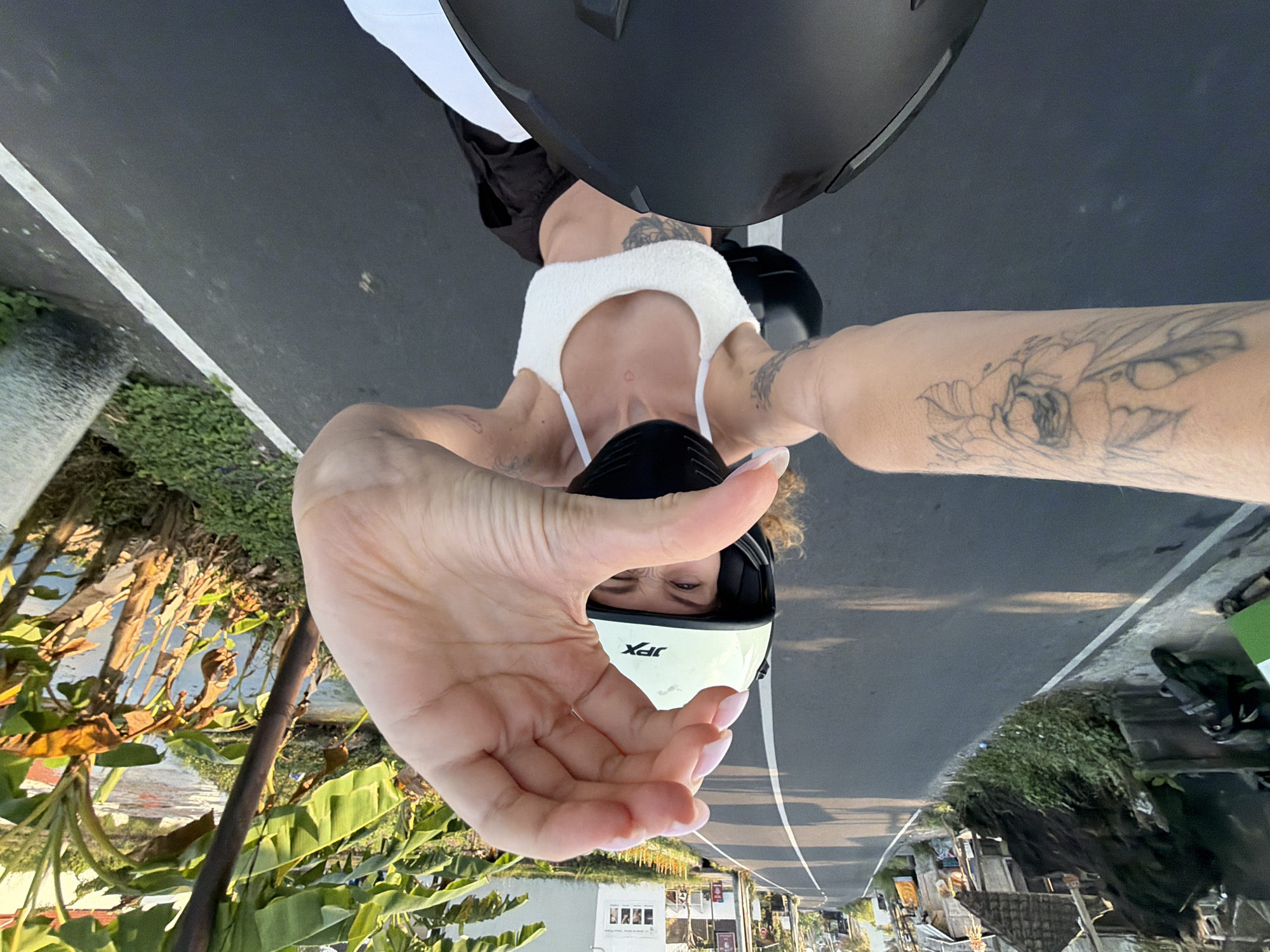 The image captures a first-person perspective of a woman riding a motorcycle on a paved road in the city of Mengwi, Indonesia. The road is bordered by lush greenery and small buildings on the left side, and dense tropical foliage, including banana trees, on the right. The woman is wearing a white cropped top, black pants, a black face mask, and a light-colored helmet. Her left arm is extended towards the camera, with her hand forming a heart shape. Tattoos are visible on her left arm and torso. In the foreground, the front of another motorcycle helmet is visible, suggesting the image is a selfie taken by the rider. The scene is illuminated by natural sunlight, indicating it is daytime.