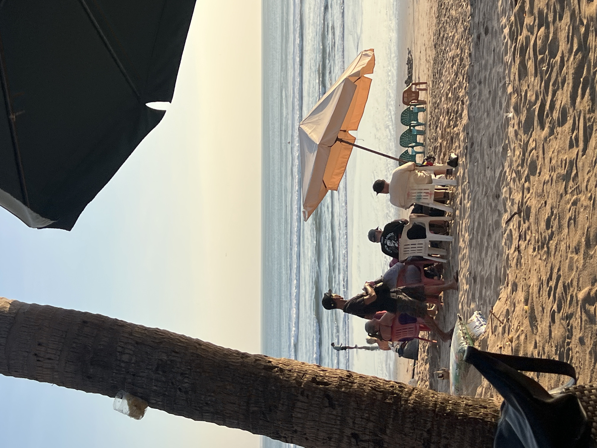 In Kuta, Indonesia, a beach scene unfolds with a group of people gathered on the sand. In the foreground, a portion of a palm tree trunk is visible on the left, with a plastic bag containing what appears to be food scraps attached to it. In the middle ground, several individuals are seated in chairs, some under an orange umbrella, facing the ocean. One person stands near the palm tree, wearing a dark t-shirt and a baseball cap. The ocean stretches out in the background, with waves breaking on the shore. A larger, dark umbrella is visible in the upper right corner of the frame, casting a shadow. The sand is golden, and the sky is a soft, clear blue, transitioning to a pale yellow near the horizon.