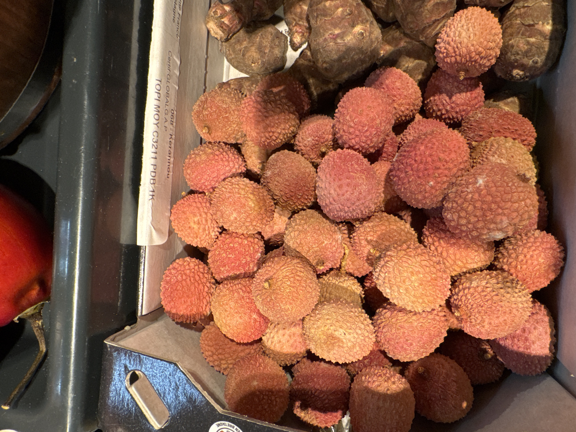 The image shows a close-up, overhead view of a cardboard container filled with lychee fruits. The lychees are predominantly red and pink, with a rough, bumpy texture. Some are clustered together, while others are more spread out. In the background, partially visible, are what appear to be root vegetables, possibly ginger or taro, with a dark brown, rough skin. The container itself is made of light brown cardboard with some printed text visible on a white label attached to the inside of the lid. The lighting is somewhat harsh, creating shadows on the fruits. The scene is set indoors, likely in a market or grocery store environment, given the packaging and arrangement of produce. The location is specified as Höhenkirchen-Siegertsbrunn, Germany. There are no people or actions depicted in the image.
