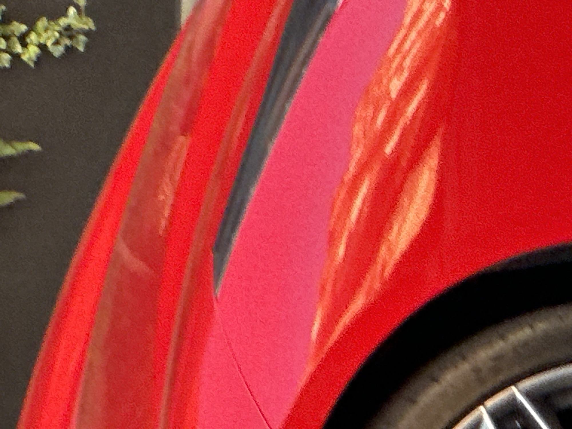 The image is a close-up, abstract shot dominated by the vibrant red bodywork of a car. It captures a section of the car's fender or side panel, showcasing its curved surface and glossy finish. A dark, possibly black, stripe or trim piece is visible, running vertically along a contour in the middle-left area of the red paint.
Bright, angular reflections in shades of orange, yellow, and white streak across the red surface, indicating strong illumination, likely from direct sunlight during the daytime. These reflections highlight the car's shape and suggest the presence of nearby structures or light sources.
In the bottom right corner, a portion of a car wheel is visible, including a black tire and a silver, multi-spoke metallic rim. The area directly above the tire, underneath the car's red arch, is cast in deep shadow.
The background to the top-left of the car features a dark, possibly grey or black, textured surface, resembling a wall or building. Sparse green foliage, appearing as small leaves or branches, is visible against this dark background in the upper left corner. The image has a slightly grainy texture overall. There are no people, explicit activities, or visible text. The precise make or model of the car is not identifiable.