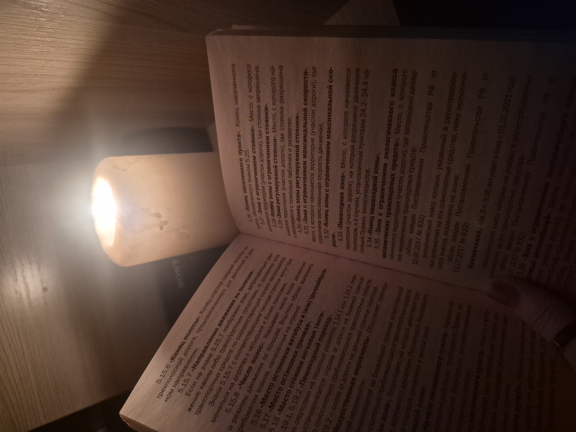 The image captures a close-up view of an open book or document with Russian text, illuminated by a warm light source, possibly a lamp or candle, positioned to the right. The book rests on a surface that appears to be wood, visible on the left side of the frame. A "SONY" logo is faintly visible on an object near the book.
The visible text in the document details various traffic rules and regulations, specifically discussing road signs and zones. Examples of sections include "Конец полосы" (End of lane), "Направление движения по полосам" (Direction of movement by lanes), "Место остановки автобуса и (или) троллейбуса" (Bus and/or trolleybus stop), "Пешеходный переход" (Pedestrian crossing), "Искусственная неровность" (Speed bump), "Начало населенного пункта" (Beginning of a populated area), "Зона с ограничением стоянки" (No parking zone), "Зона с ограничением максимальной скорости" (Speed limit zone), "Пешеходная зона" (Pedestrian zone), and "Зона с ограничением экологического класса механических транспортных средств" (Zone with restriction on the ecological class of mechanical vehicles). The text also references "Постановление Правительства РФ" (Decree of the Government of the Russian Federation) and specific dates like 12.07.2017 and 01.07.2021, indicating legal or official documentation. The overall setting, likely indoors, suggests a moment of reading or studying, possibly in the evening given the reliance on artificial light.