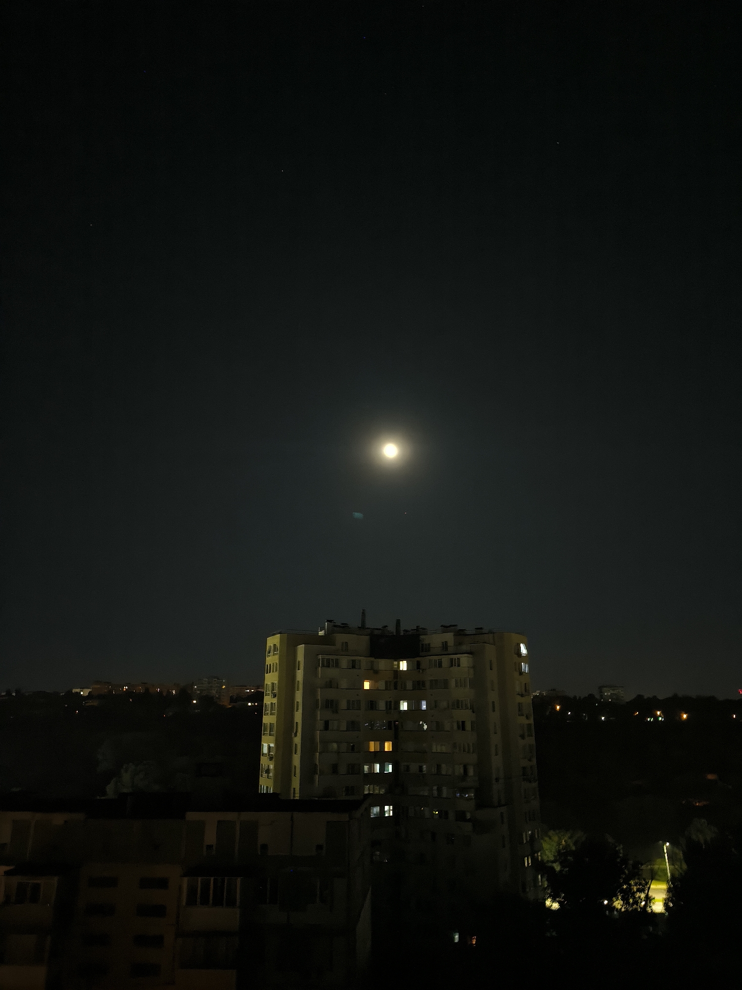 This is a vertical, eye-level, nighttime photograph taken from an elevated vantage point, looking out over a residential area in Chișinău, Moldova. The sky is a deep, dark blue with a bright, full moon positioned just above the center of the frame. The moon has a strong, luminous glow, with a faint lens flare visible below it. A few distant stars are discernible in the dark sky.
In the mid-ground, a multi-story, light yellow apartment building dominates the center of the image. It has a somewhat unusual, curved facade and numerous windows, many of which are illuminated with a warm, yellowish light, suggesting occupancy. The building's roofline features several visible antennas and structures.
In the foreground, the top of another, lower apartment building is visible, appearing darker and less illuminated than the central structure. Its windows are mostly dark, with a few exceptions showing faint lights. The architecture suggests a Soviet-era housing block.
The background consists of a dark, silhouetted landscape of trees and other buildings spread across the horizon. Scattered lights from other distant structures punctuate the darkness. A street lamp with a bright light illuminating the ground is visible on the right side of the frame, partially obscured by trees. The overall scene conveys a sense of urban nighttime tranquility, with the illuminated windows providing a human element against the vastness of the dark sky and the moon.