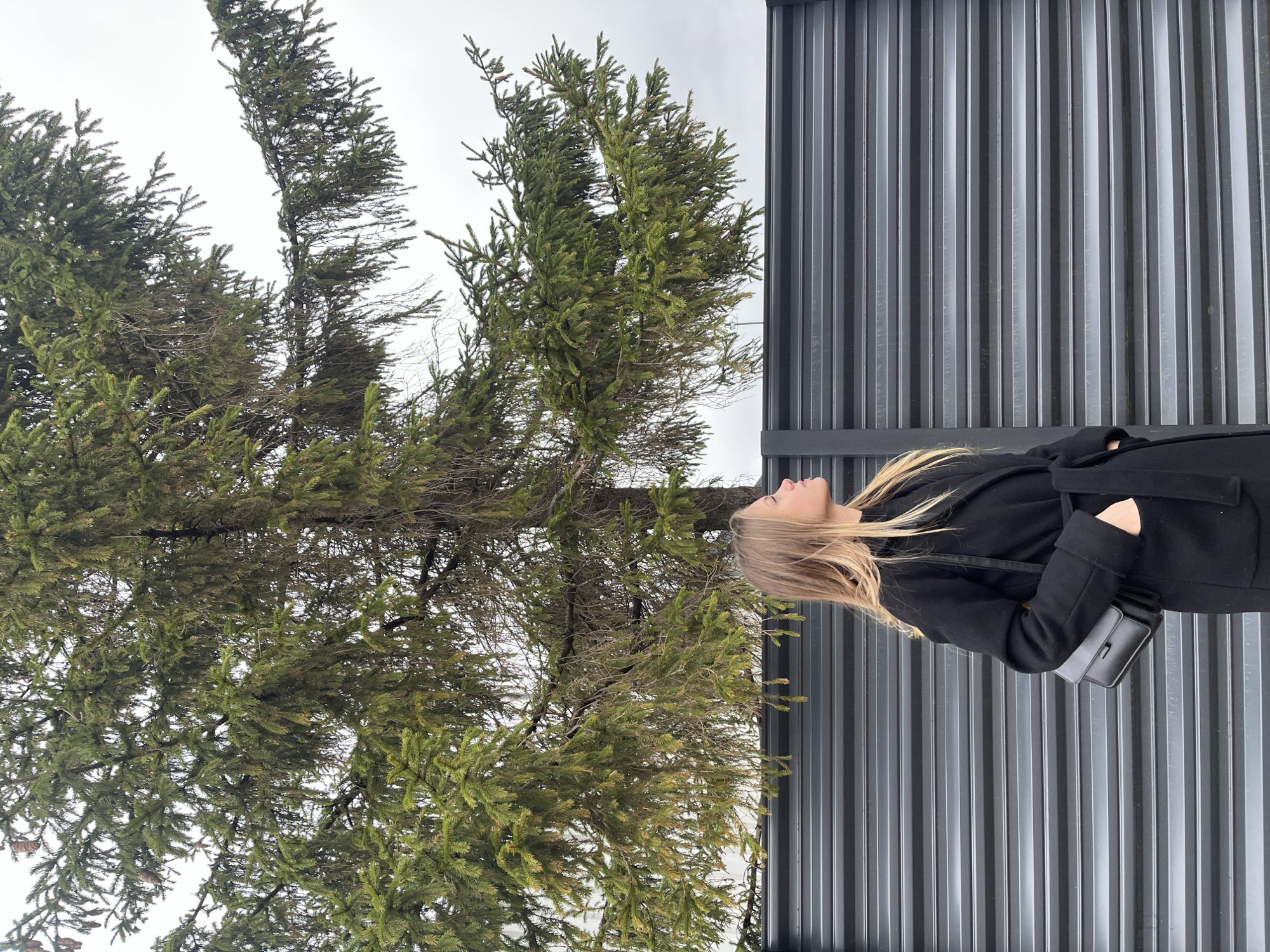 A woman in a black coat stands next to a dark, corrugated metal wall, looking up at a large evergreen tree against a pale, overcast sky. The tree dominates the left side of the image, its branches reaching towards the upper left corner. The location is Фоки, Russia.