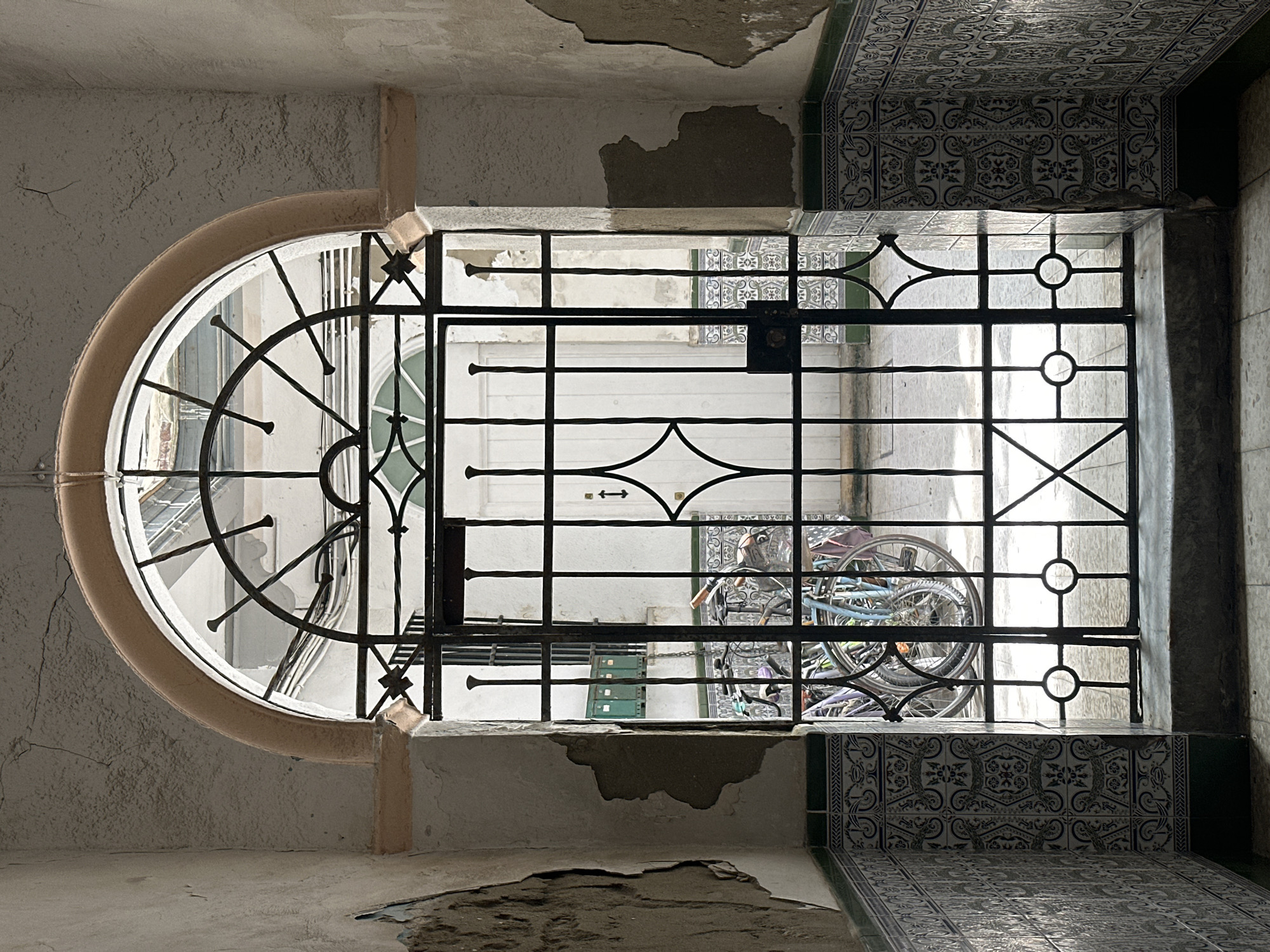This is an exterior, eye-level, medium shot of an ornate wrought iron gate and archway leading into a courtyard in Cádiz, Spain. The gate is dark and features a diamond pattern in the center and circular accents at the bottom. Beyond the gate, several bicycles are parked in the courtyard, which has a tiled wall and a white door. The surrounding hallway has cracked, textured walls with peeling paint and decorative tiled wainscoting. The archway above the gate is decorated with a light pink, stucco-like material. The scene is lit by natural light, casting shadows through the gate.