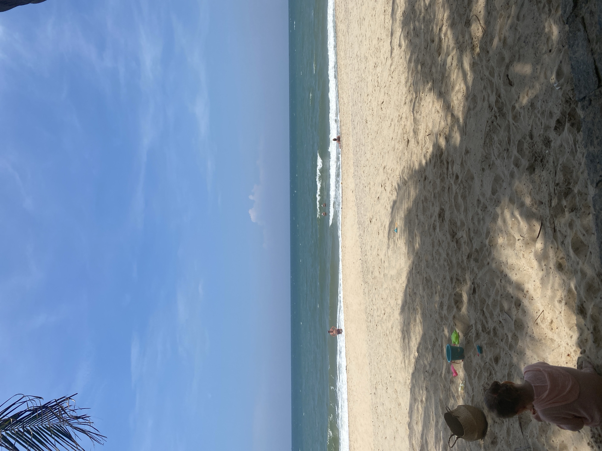 This is a coastal scene at Phu Thuy Ward, Vietnam. The view includes a clear blue sky, a turquoise sea with gentle waves, and a sandy beach. Three individuals are visible in the sea, with one closer to the shore. On the beach, there is beach equipment, including a bucket. A person is playing on the beach. The lighting is bright, casting shadows on the sand. A palm tree is visible in the bottom left corner.