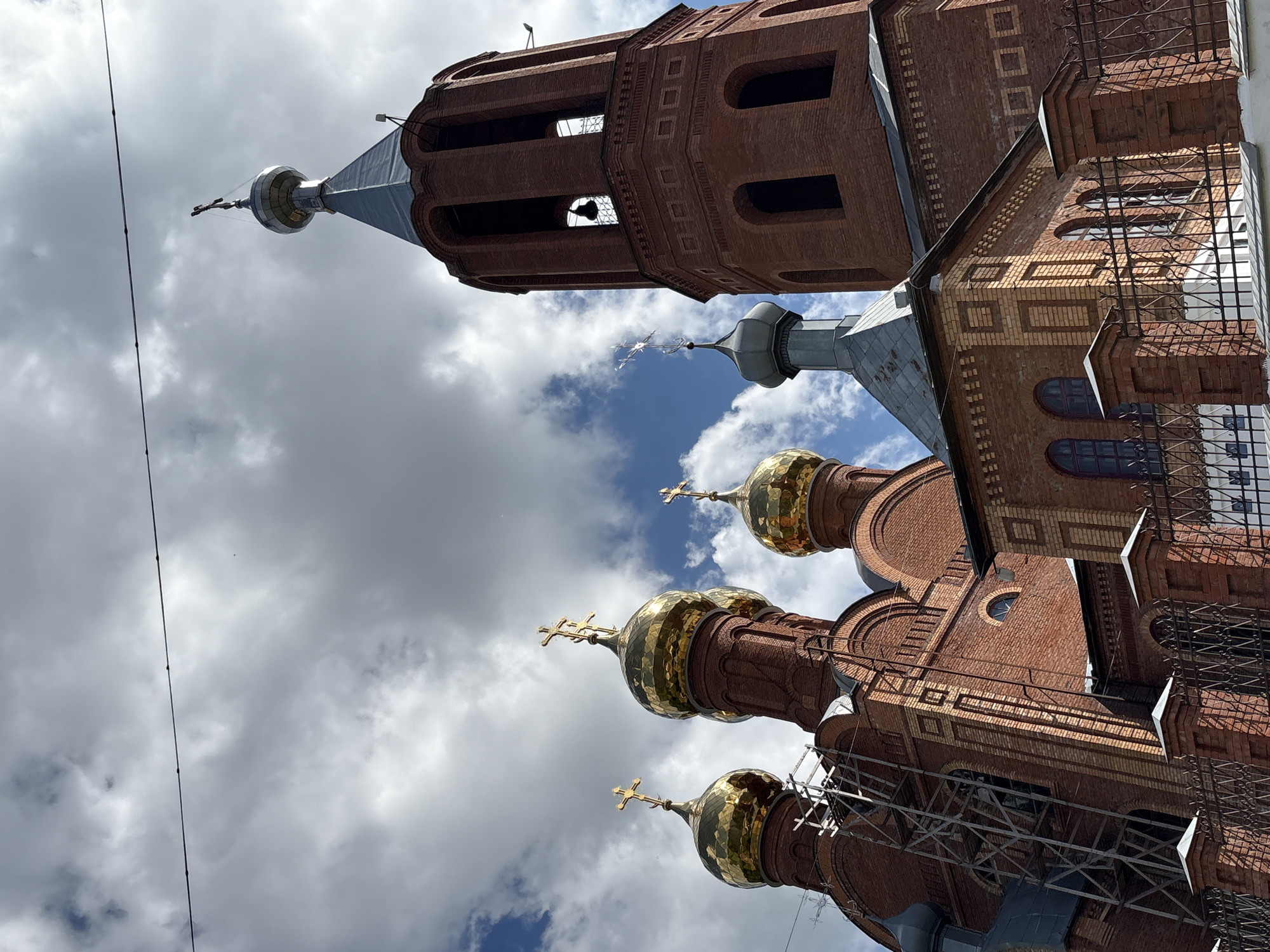The image shows a red brick Orthodox church in Kirovo-Chepetsk, Russia. The church has several golden domes topped with crosses and a tall bell tower with an arched belfry. There is scaffolding on one side of the church, suggesting ongoing construction or renovation. The sky is partly cloudy with some blue visible. A black wire runs horizontally across the top of the frame.