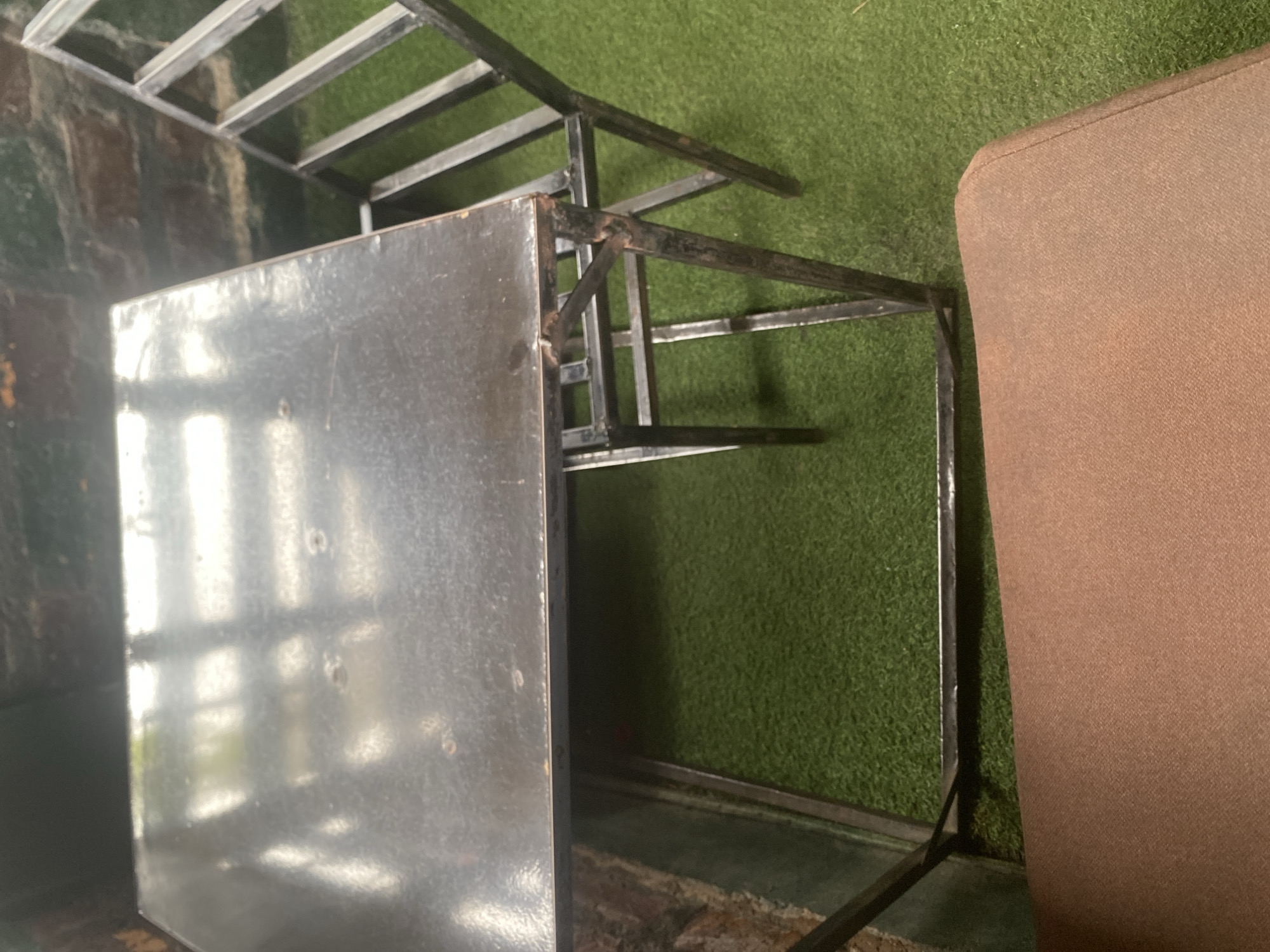 The image shows a close-up, angled view of a metal table and a portion of a brown upholstered chair. The table has a dark, reflective surface that shows reflections of overhead lighting and possibly some greenery. The metal frame of the table is visible, with visible welds and some signs of rust. The chair is upholstered in a coarse, brown fabric. The background consists of a green, artificial turf-like surface. The overall impression is that of an outdoor seating area, possibly a patio or beer garden, given the artificial turf. The lighting suggests it is daytime, though the specific time is not discernible. There are no people visible in the image, and no text is present. The scene appears to be a static arrangement of furniture. The location context provided is Jalingo, Nigeria, which does not offer specific visual cues within the image itself.