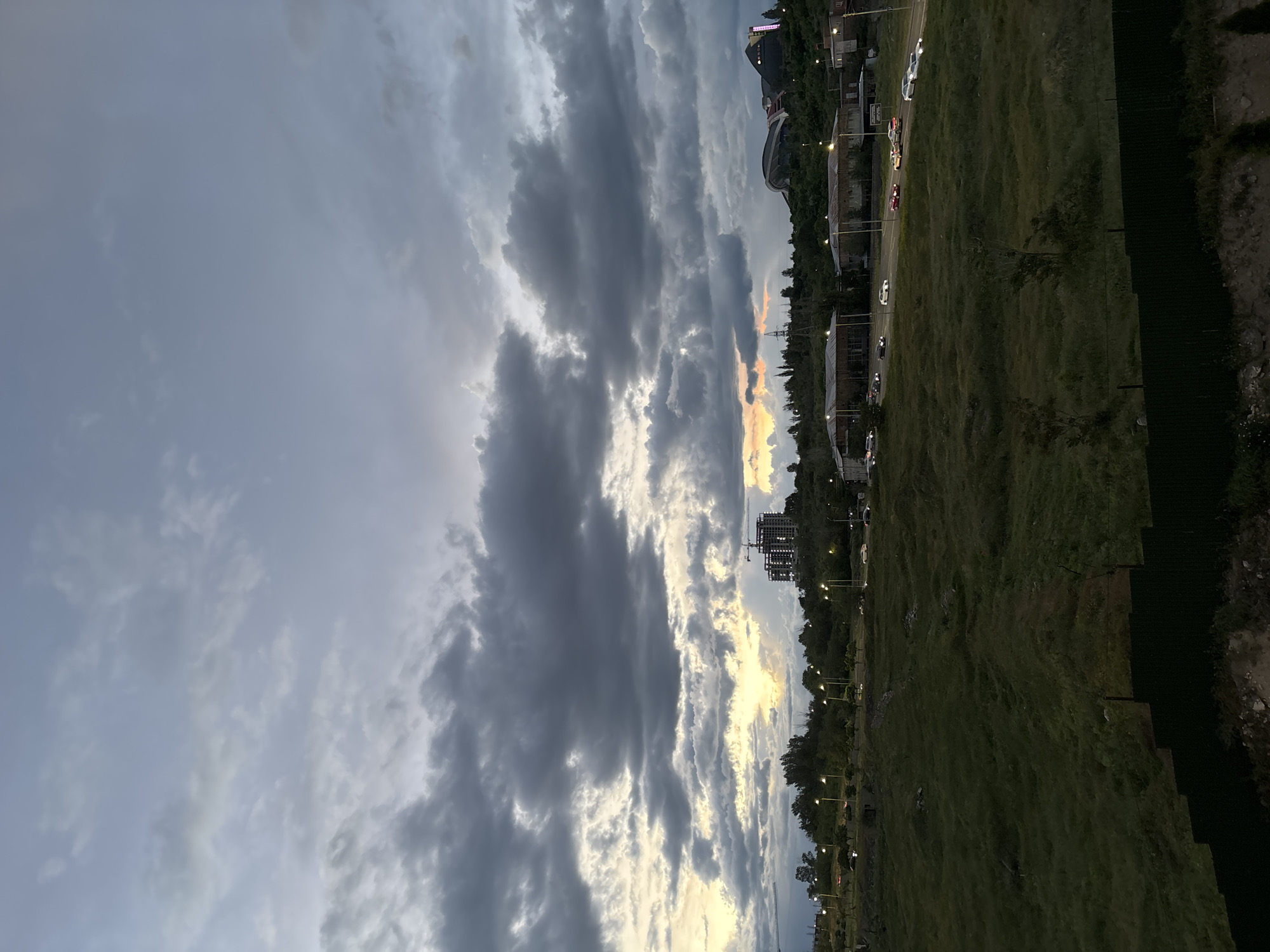 The image depicts a scene at dusk in Yerevan, Armenia. The sky is dominated by heavy, layered clouds, varying in shades of gray and white. Patches of orange and yellow are visible between the clouds.
Below the sky, a green field stretches along the right side of the image. Along the edge of the field, there are street lights and trees. In the background, there are buildings, including a large stadium with illuminated features. Cars are visible on the road along the building. A construction site with a crane is visible among the buildings.