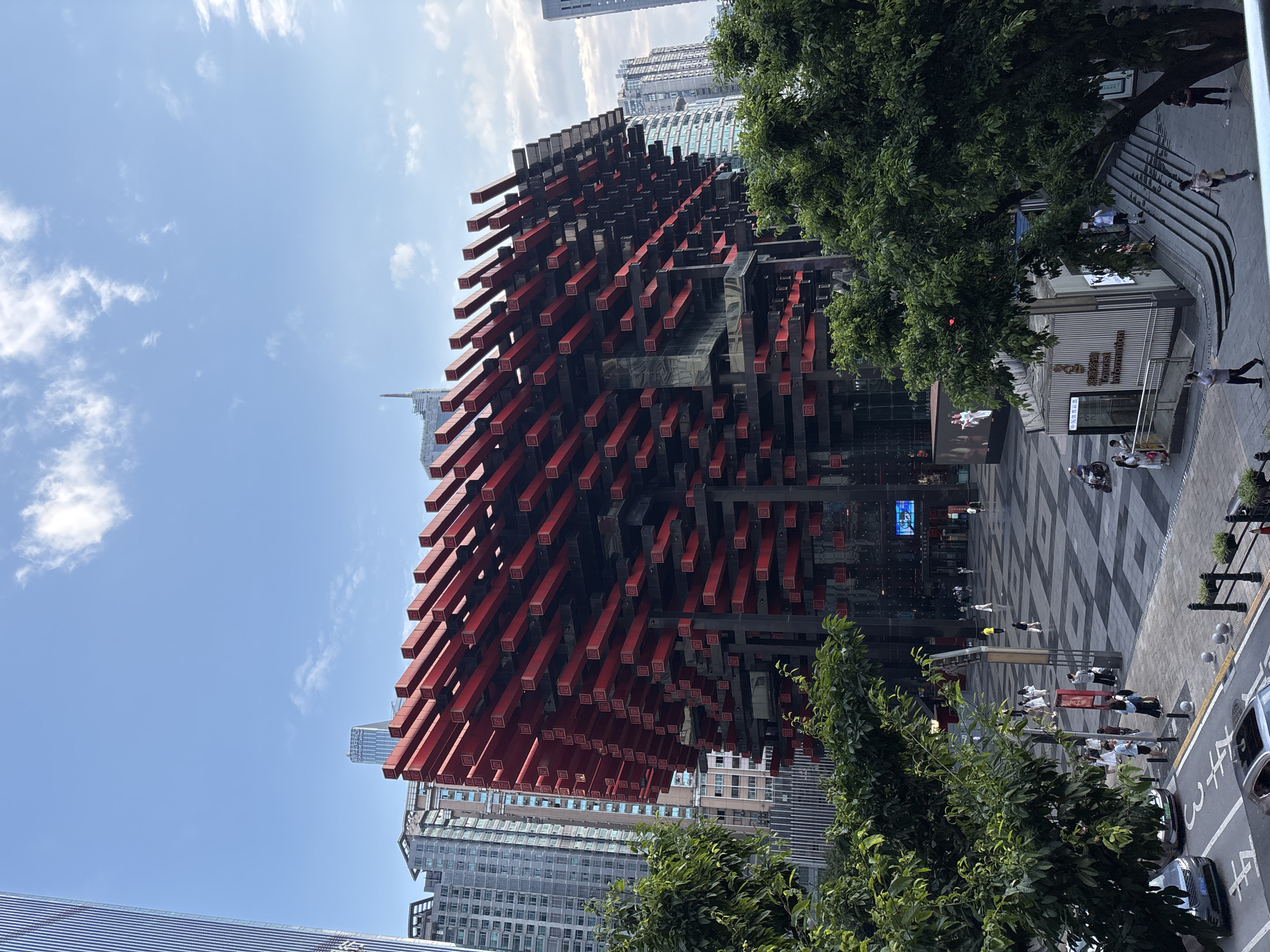 The image captures a street-level view of a modern architectural marvel in the Yuzhong District, China. Dominating the center is a building with a striking facade characterized by numerous protruding, rectangular red elements, creating a layered, three-dimensional effect. These red elements are stacked and staggered against a dark background, forming a dynamic, almost crystalline structure.
Surrounding this central building are contemporary high-rise structures, typical of an urban cityscape. Lush green trees are strategically placed in the foreground, partially obscuring the lower levels of the buildings and the street scene below.
On the ground level, a paved plaza features a geometric pattern. Several individuals are walking across the plaza, some in small groups, suggesting a public space with pedestrian activity. There are also several parked cars visible on the adjacent street, including a silver sedan and a darker SUV.
A small kiosk-like structure with signage, likely an information booth, is situated near the base of the building, with people interacting in its vicinity. The sky above is a clear blue with scattered white clouds, indicating fair weather. The overall impression is a blend of cutting-edge architecture and vibrant urban life.